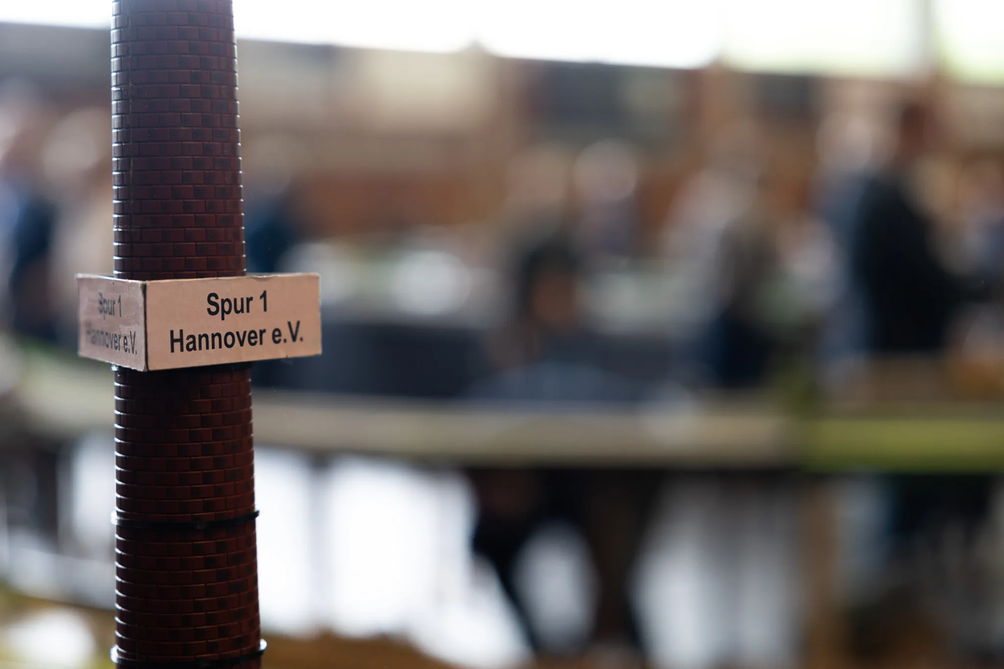 Schild „Spur 1 Hannover e.V.“ an einem Pfosten vor verschwommenem Hintergrund von Besuchern einer Modellbahn-Ausstellung.