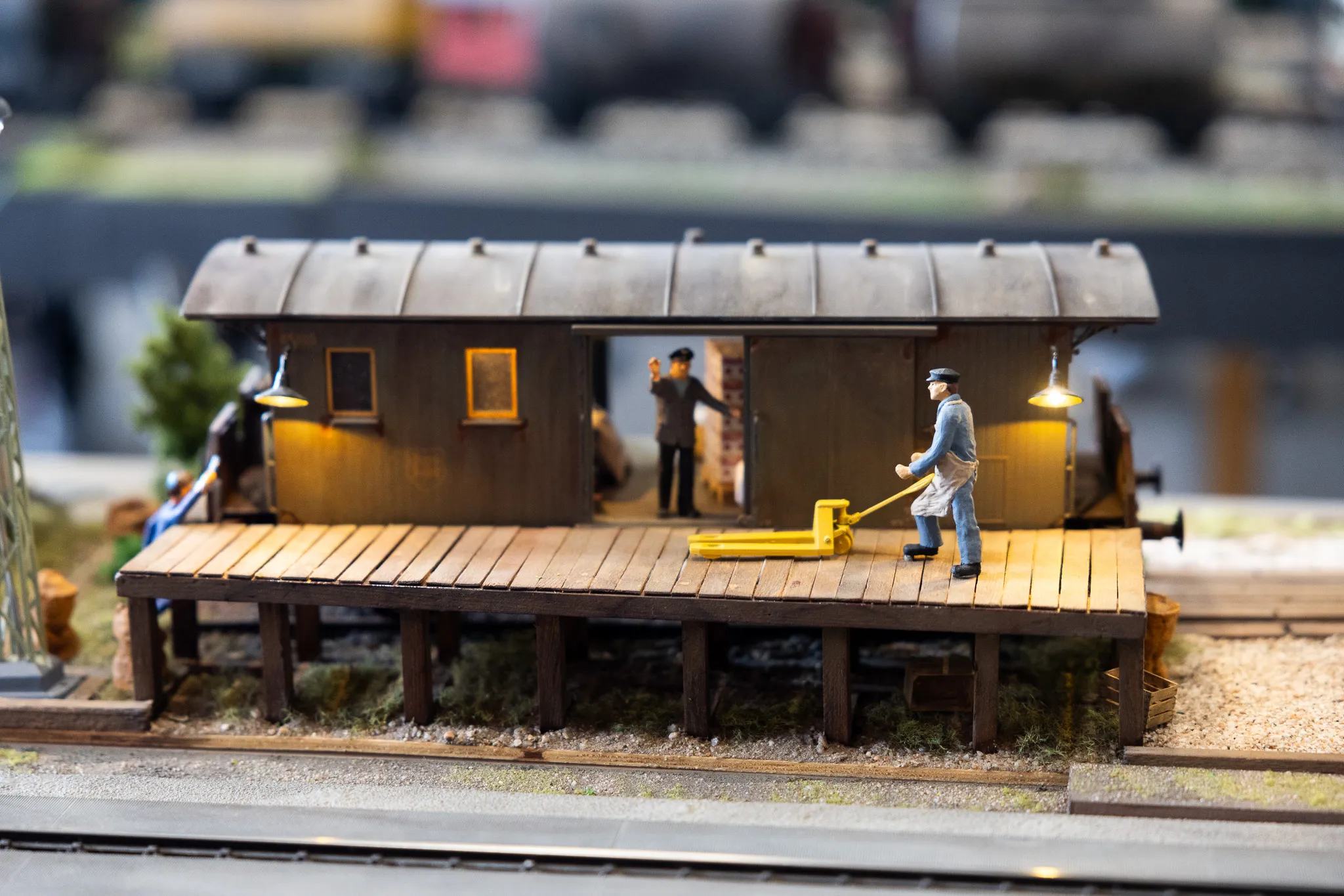 Ein Mann in Arbeitskleidung steht vor einem Modellbahnhof und schiebt eine gelbe Plattform. Im Hintergrund sind weitere Figuren und Details der Modellbahn zu sehen.