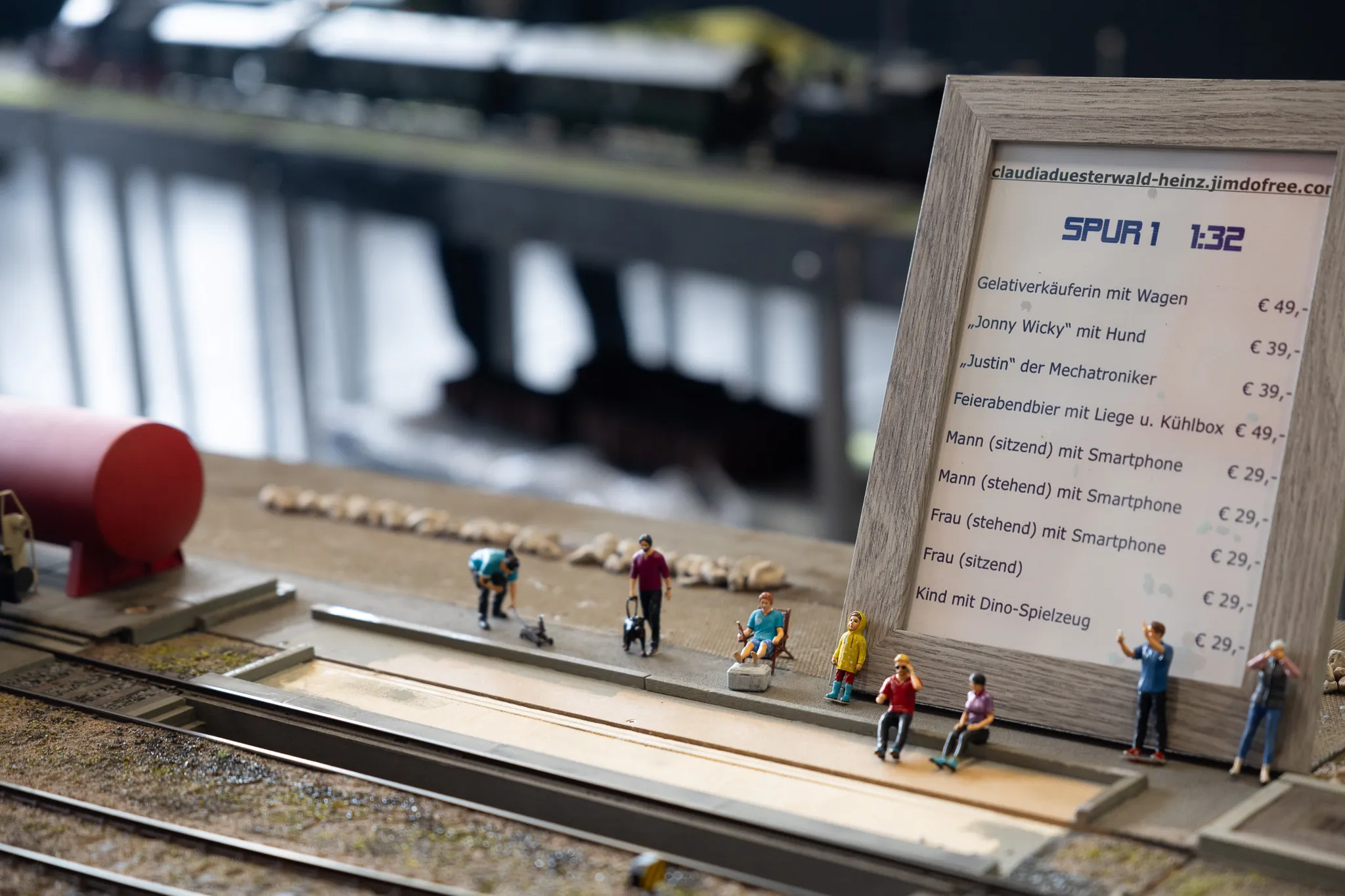 Miniaturfiguren und eine Preisliste stehen auf einer Modellbahnanlage mit Gleisen und Landschaftselementen.