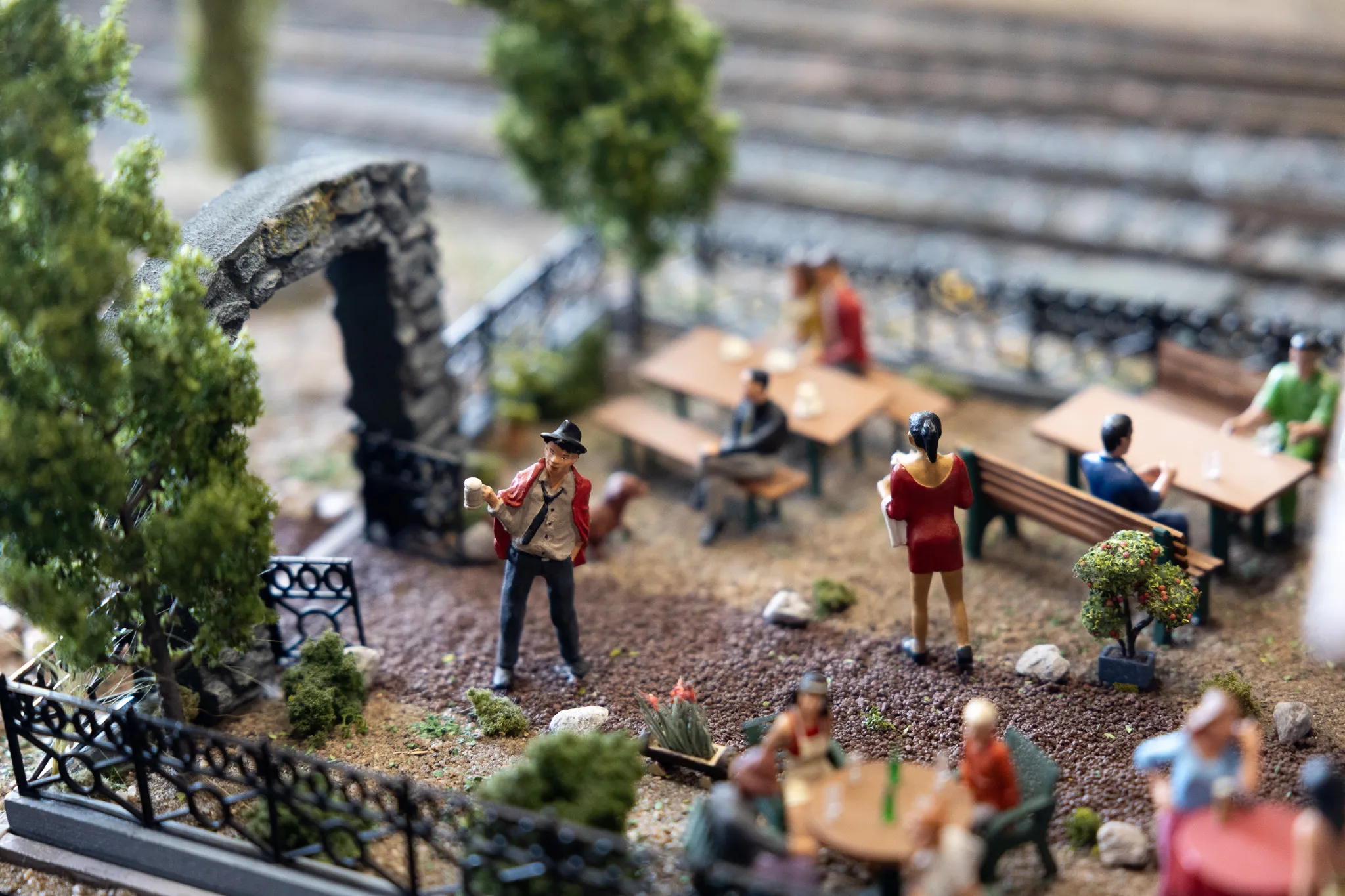 Miniaturfiguren in einem Modellbahn-Szenario zeigen Menschen in einem Biergarten vor einem Gebäude und einer Mauer.