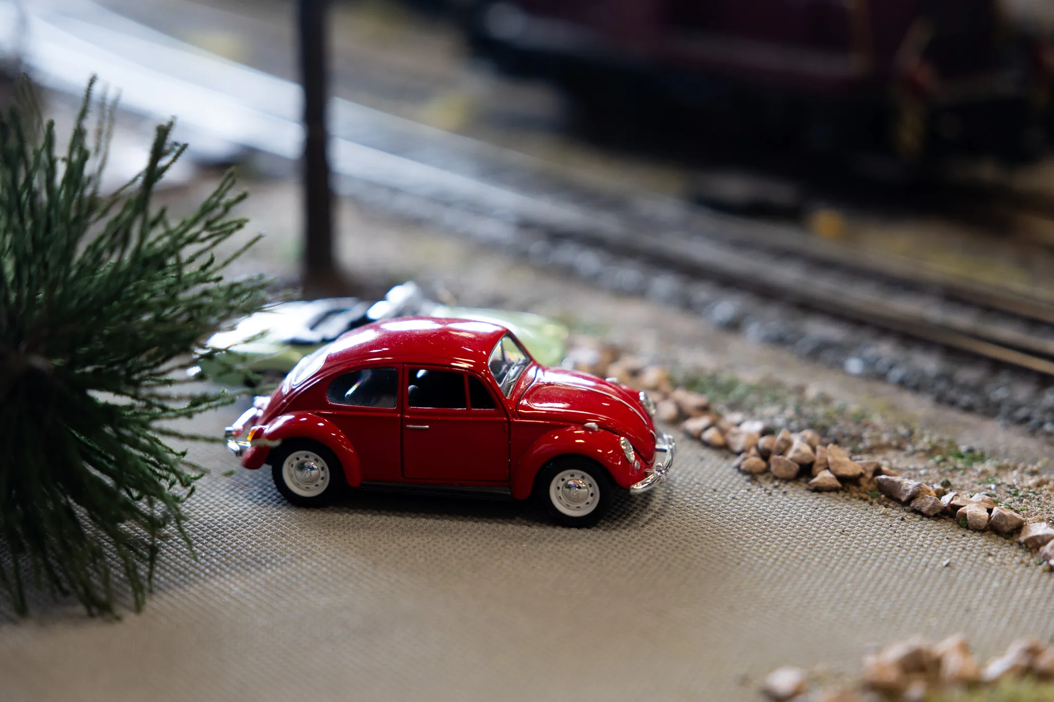 Roter Modell-Käfer fährt auf einer Modellbahnstraße neben einem kleinen Nadelbaum.
