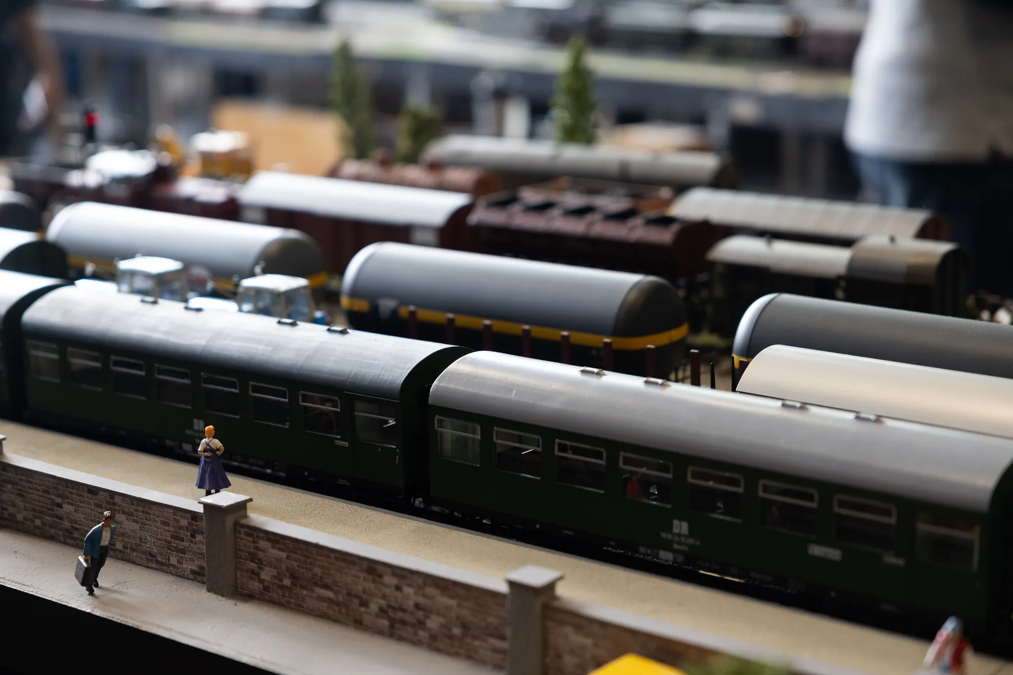 Mehrere detailreiche Modelle von Waggons stehen auf Gleisen vor einer Mauer, während eine Frau mit Hut und ein Mann am Bahnsteig stehen.