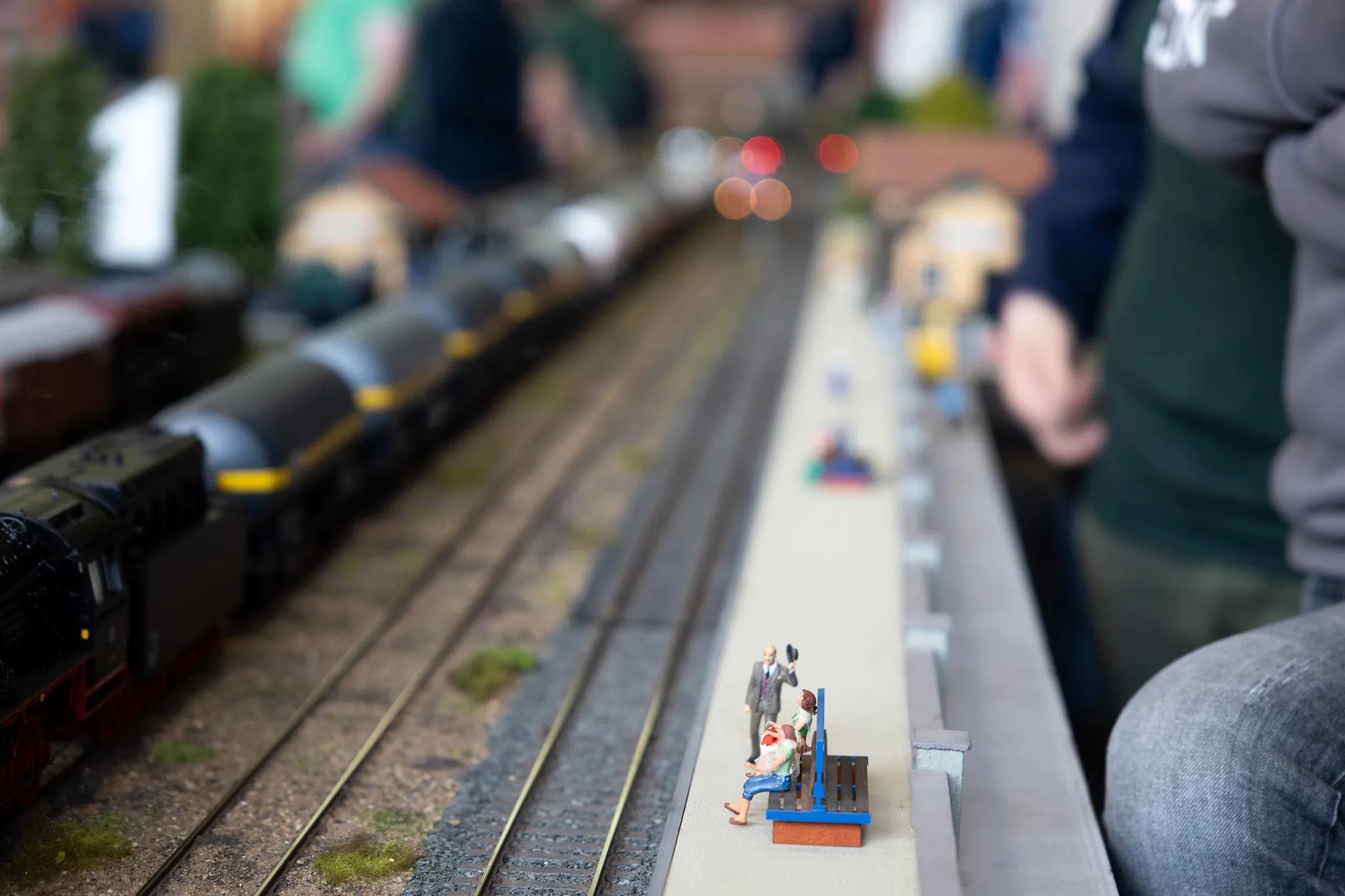 Eine rote Dampflok und Waggons stehen auf Gleisen einer Modellbahn, neben der eine kleine Gruppe von Miniaturfiguren auf einer Bank sitzt.