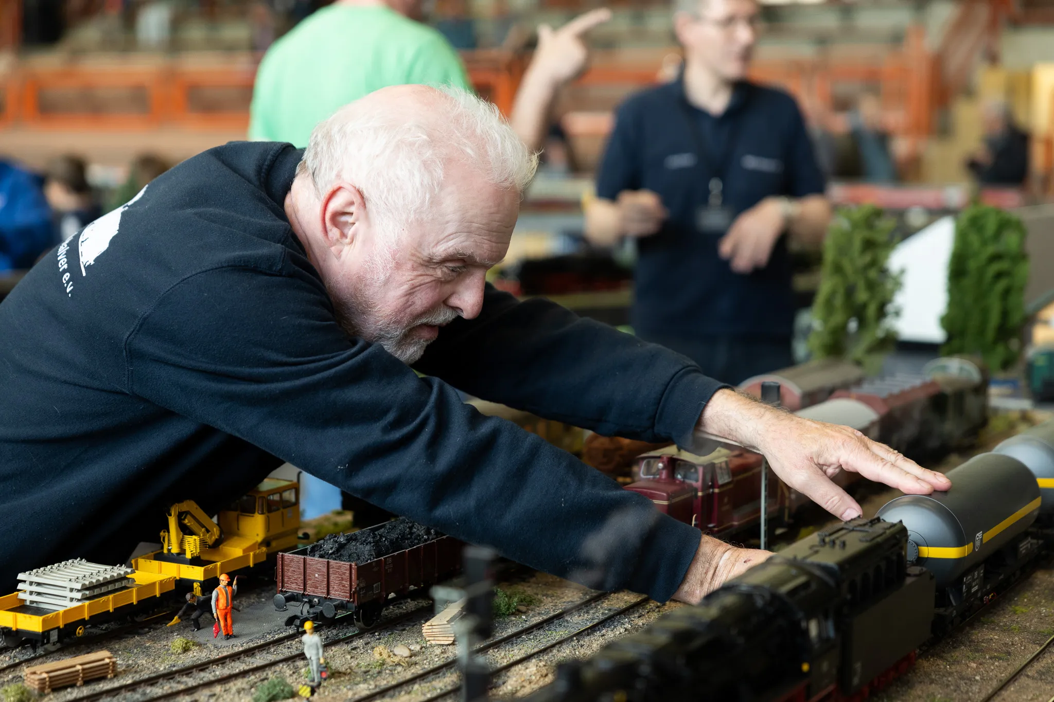 Ein älterer Mann beugt sich konzentriert über eine detailreiche Modelleisenbahnanlage mit mehreren Waggons und einer Lokomotive.