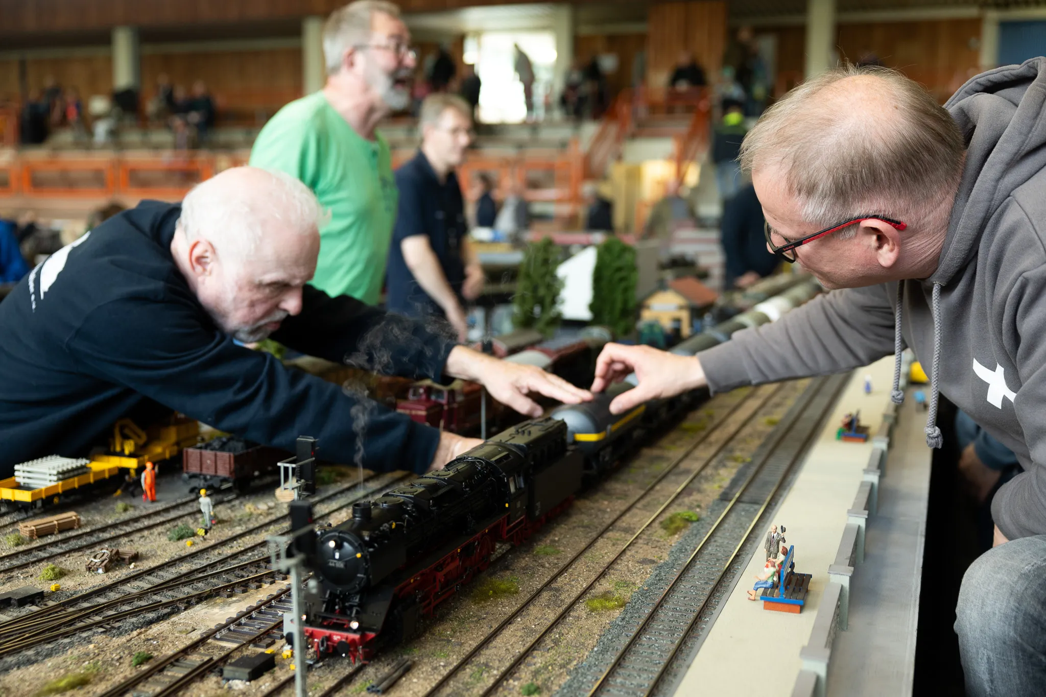 Zwei Männer betrachten eine detailreiche Modelleisenbahnanlage mit einer Dampflok und mehreren Waggons. Im Hintergrund weitere Besucher der Ausstellung.