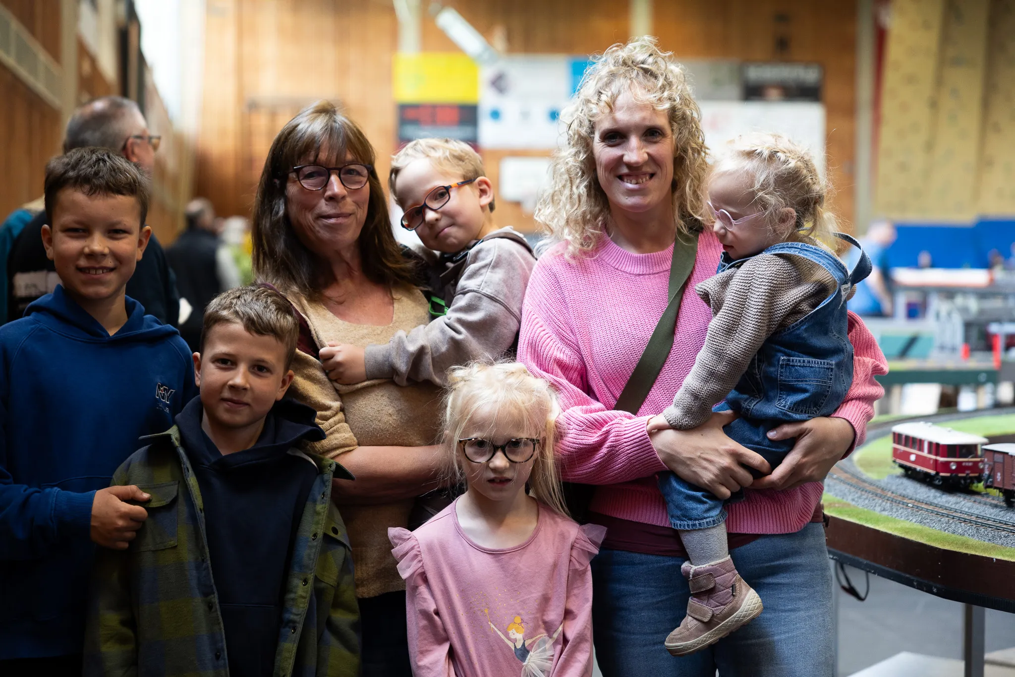 Eine Frau mit lockigem Haar hält ein Kleinkind im Arm, umgeben von vier Kindern und einer weiteren Frau. Im Hintergrund ist eine Modelleisenbahnanlage zu sehen.