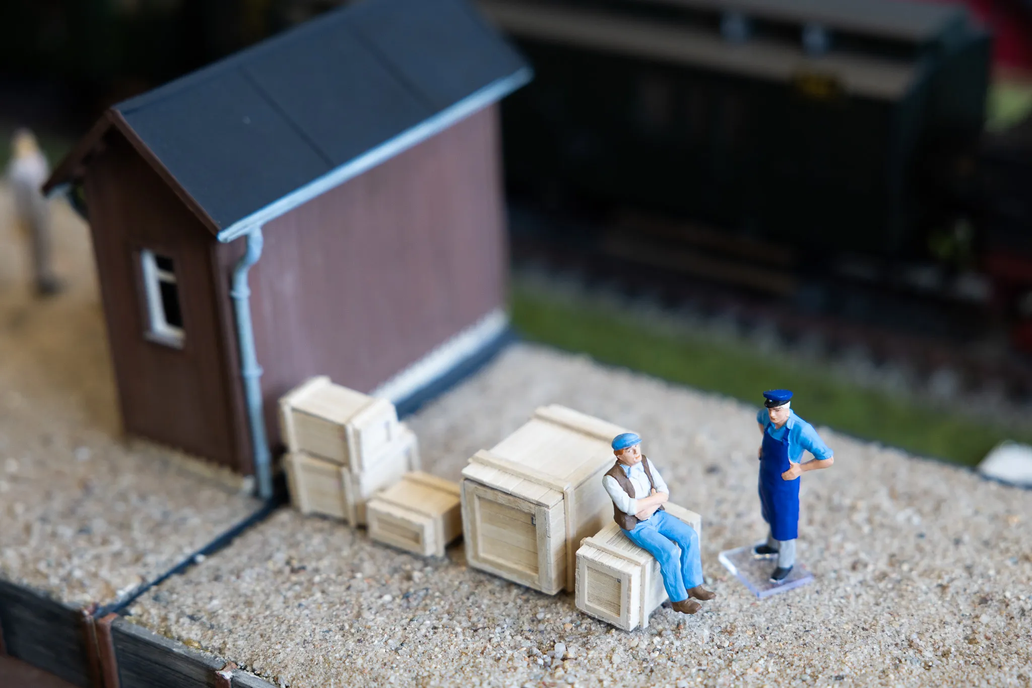 Zwei Miniaturfiguren sitzen und stehen vor einem kleinen braunen Schuppen und mehreren Holzkisten auf einem Kiesweg.