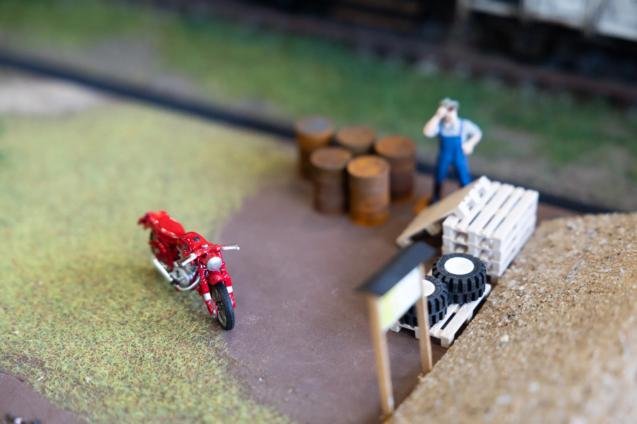 Rote Modellmotorrad neben Holzstämmen und einer Figur in Arbeitskleidung auf einer Modellbahnanlage.