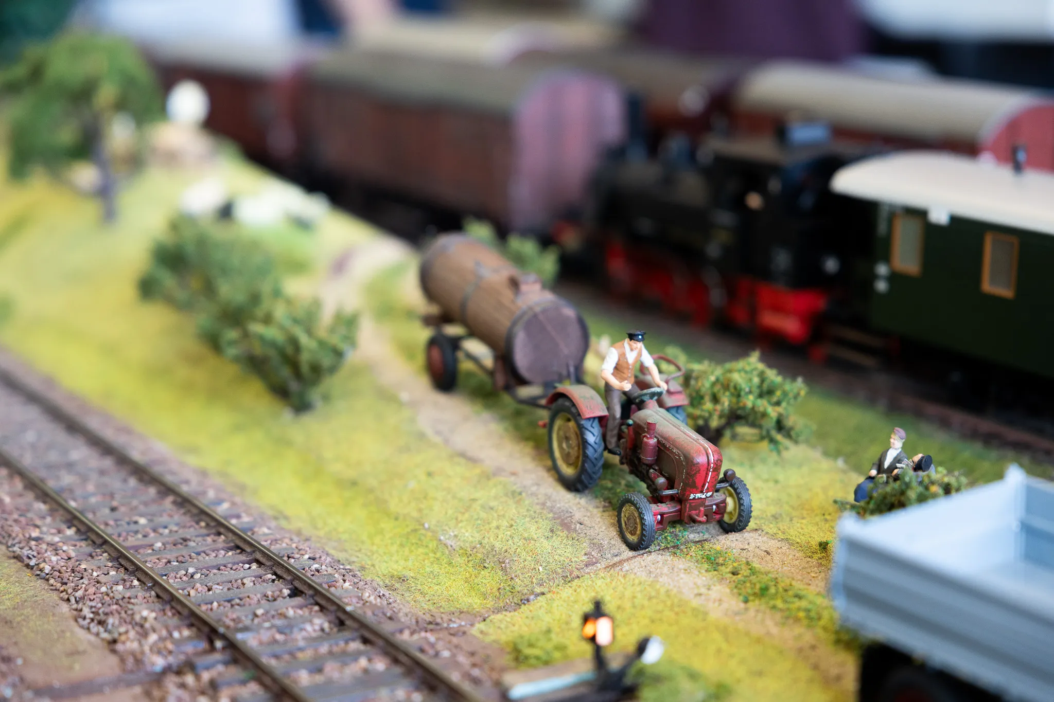 Roter Traktor mit Anhänger befördert einen großen Tank auf einer Modellbahnlandschaft entlang von Gleisen.
