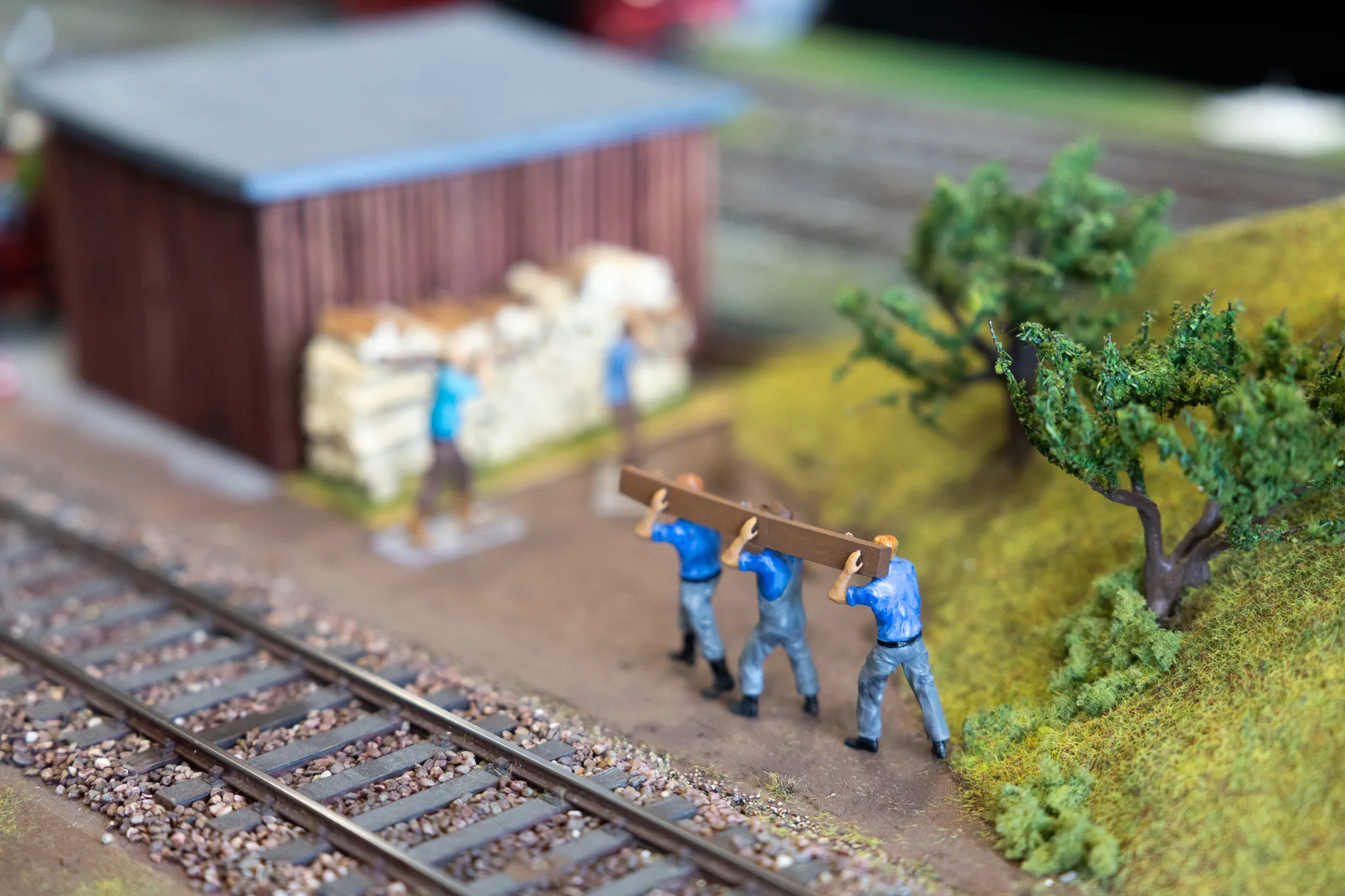 Drei Modellfiguren tragen gemeinsam ein langes Brett neben einer Modelleisenbahnstrecke an einem kleinen Gebäude vorbei.