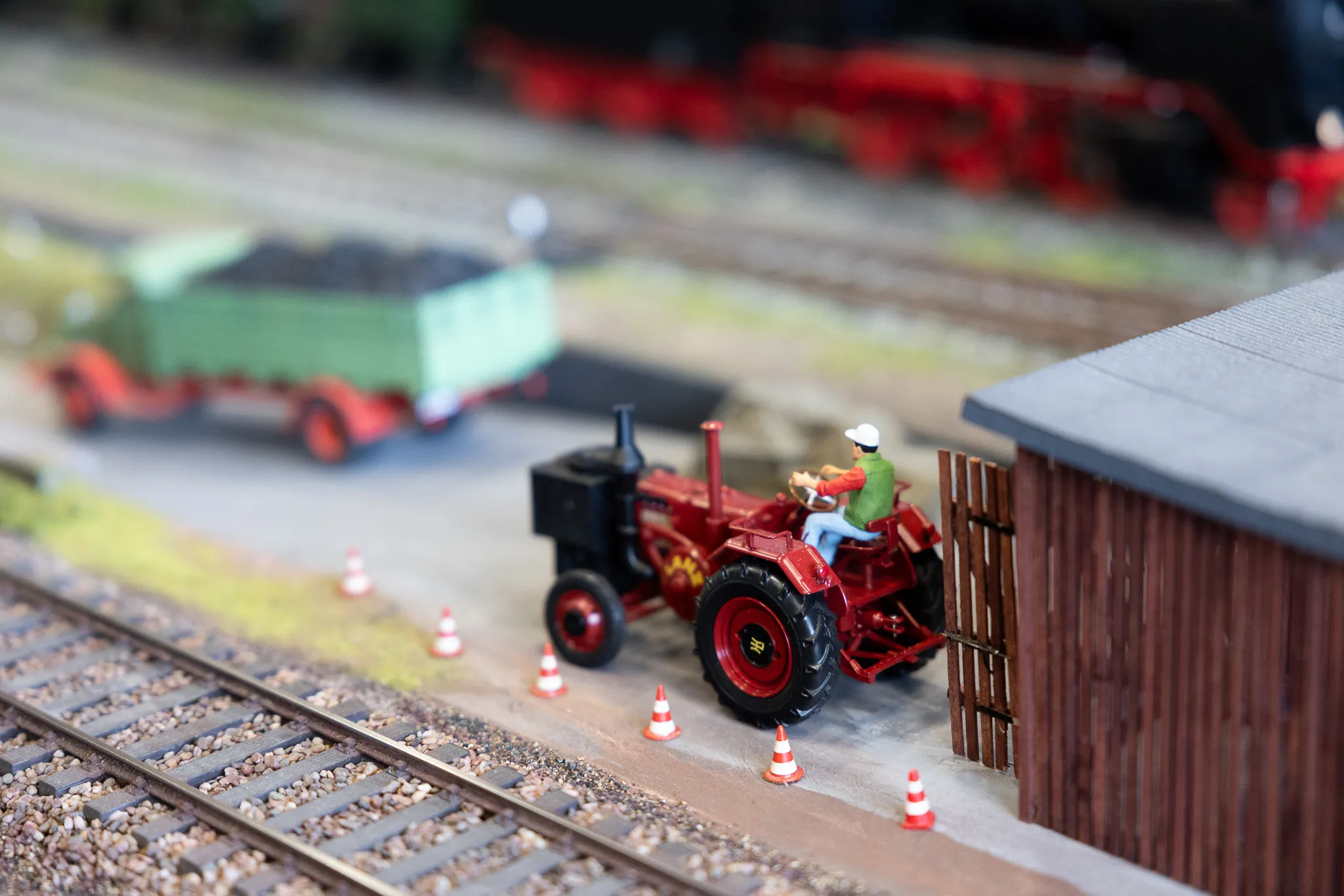 Roter Traktor mit Fahrer fährt an einer Modelleisenbahnstrecke vorbei, umgeben von Miniatur-Verkehrskegeln und einem Lagerhaus.