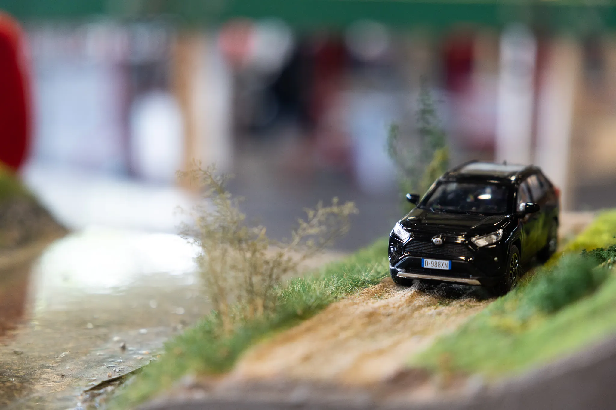 Schwarzes Modellauto auf einer Miniaturlandschaft mit Büschen und einem Flussbett, im Hintergrund verschwommene Besucher einer Ausstellung.