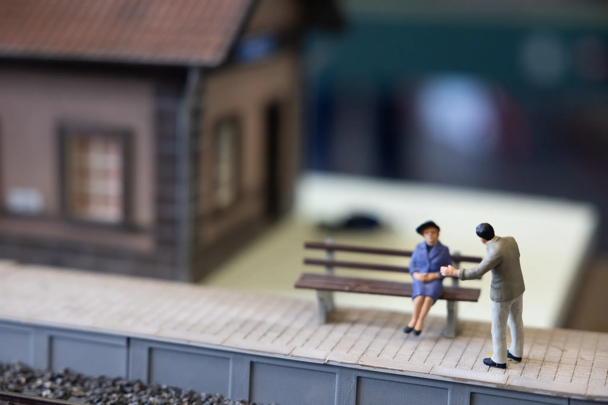 Miniaturfiguren von Mann und Frau auf einer Bank vor einem Modellbahnhof, Teil einer Modellbahnanlage.