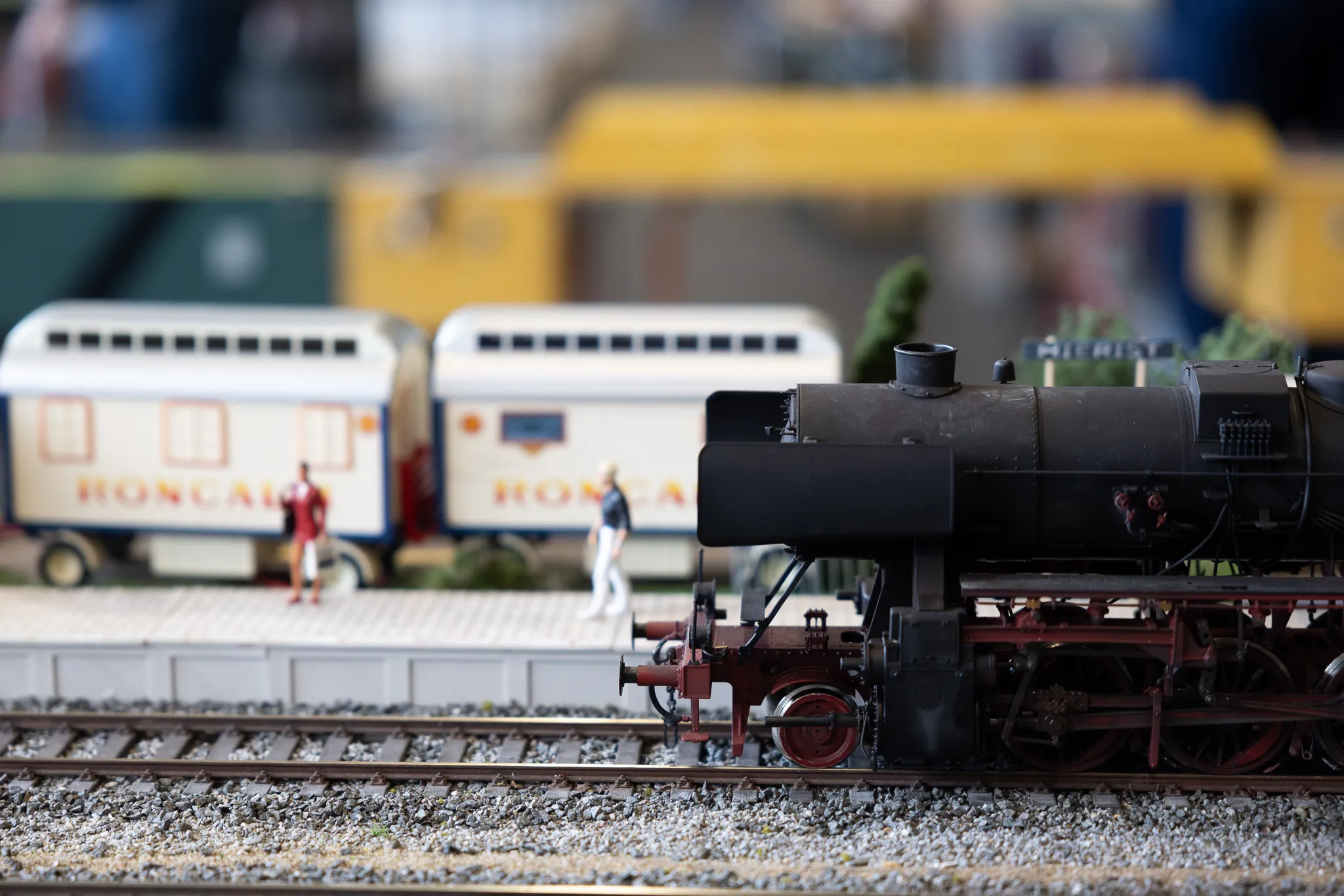 Dampflok und zwei Waggons der Modellbahn auf Gleisen, im Hintergrund verschwommene Personen.