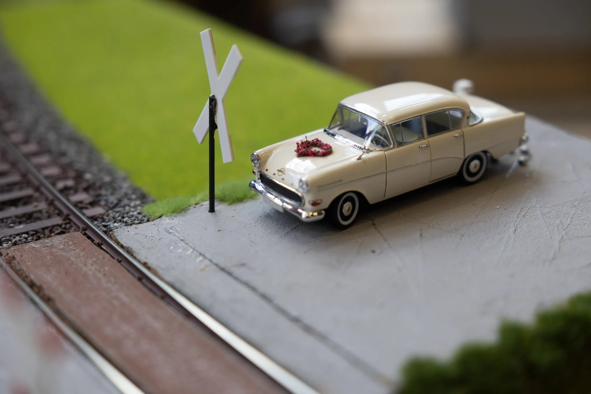 Beigefarbener Oldtimer mit Blumenschmuck auf dem Dach steht vor einem Schranken an einer Modellbahnanlage.