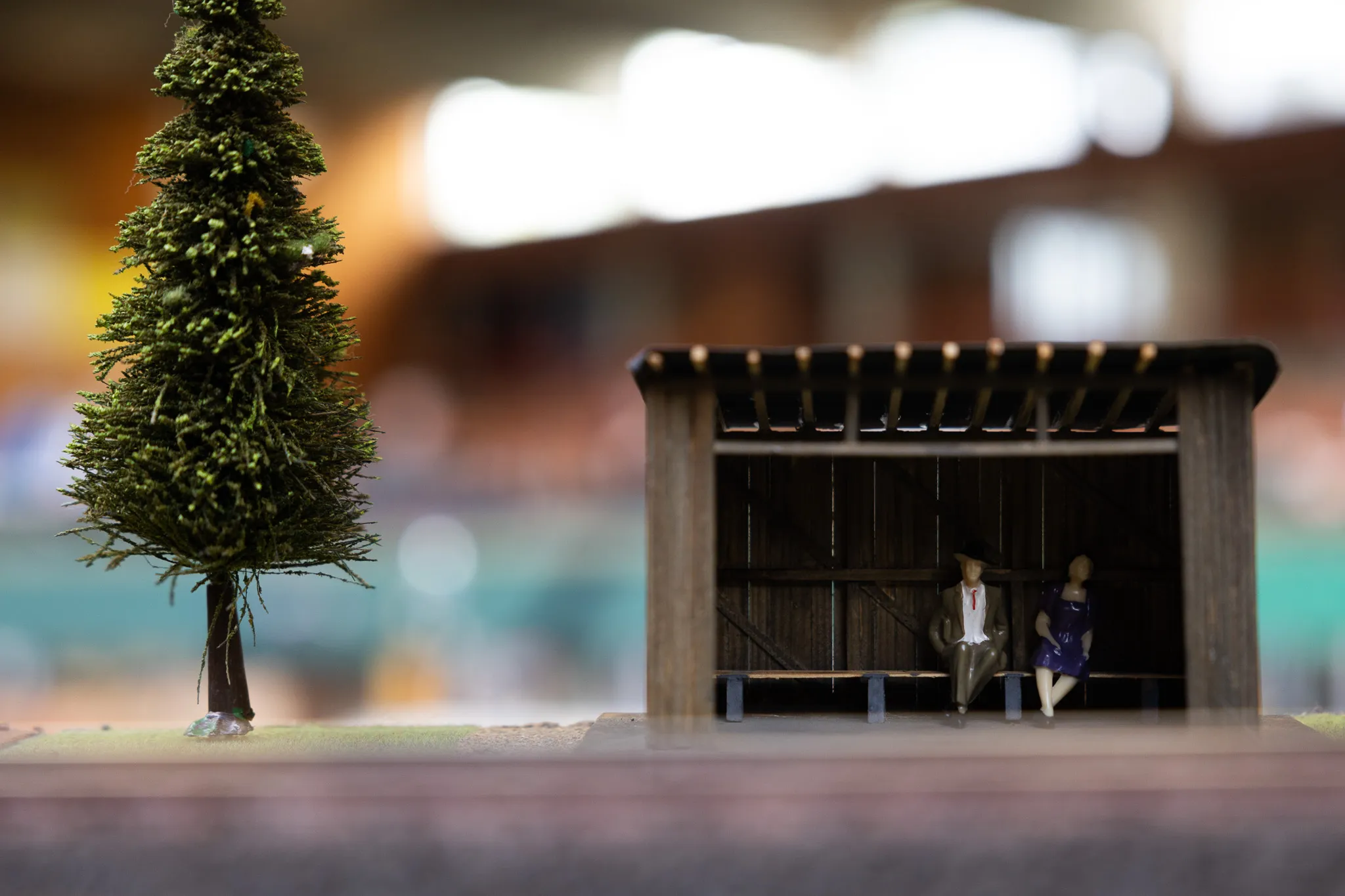 Miniatur-Buschhalte mit zwei Personen im Anzug und Kleid vor verschwommenem Hintergrund einer Modellbahn-Ausstellung.