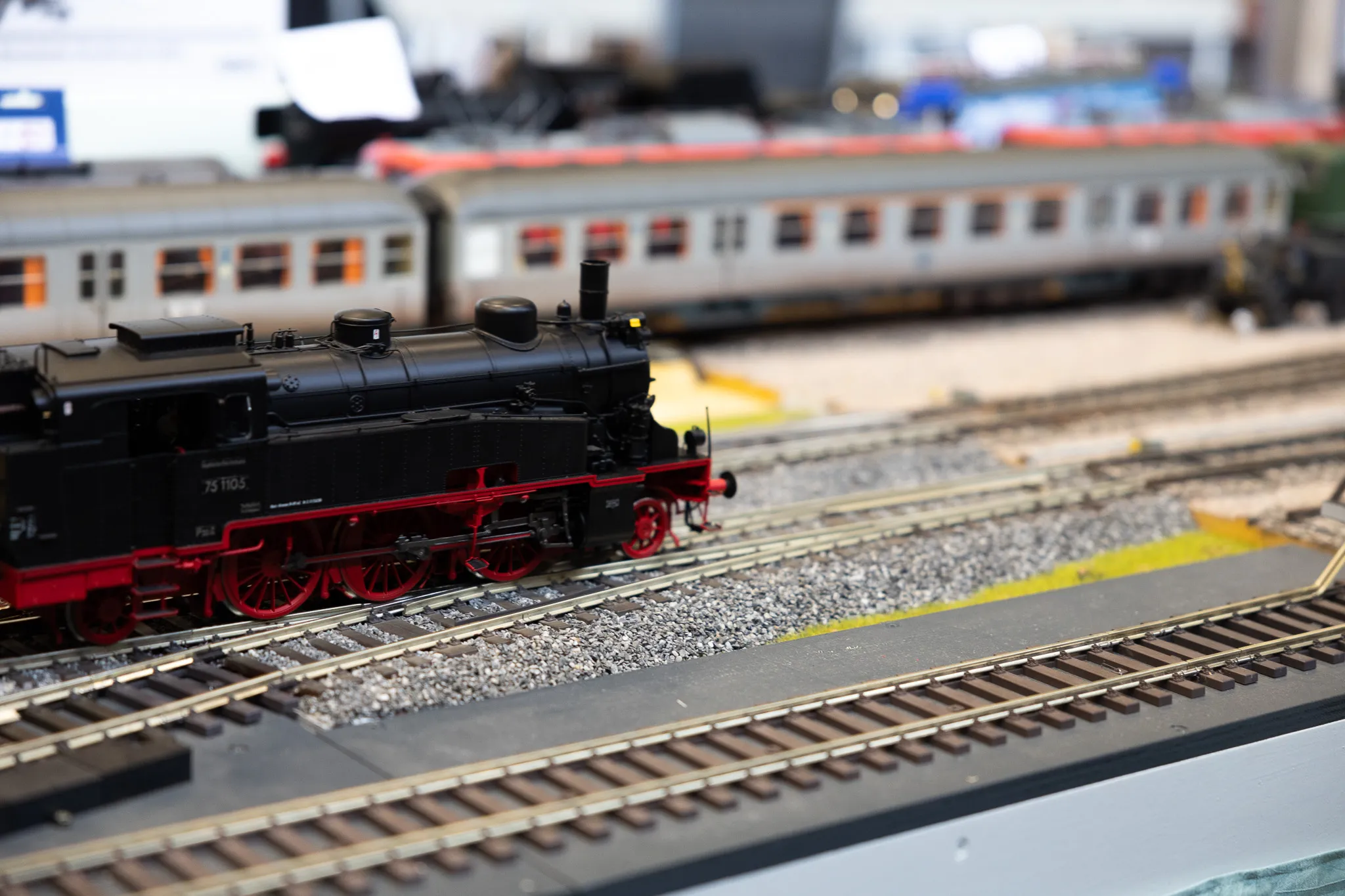 Schwarze Dampflok mit roten Rädern auf Gleisen einer Modellbahn, im Hintergrund ein beiger Waggon.