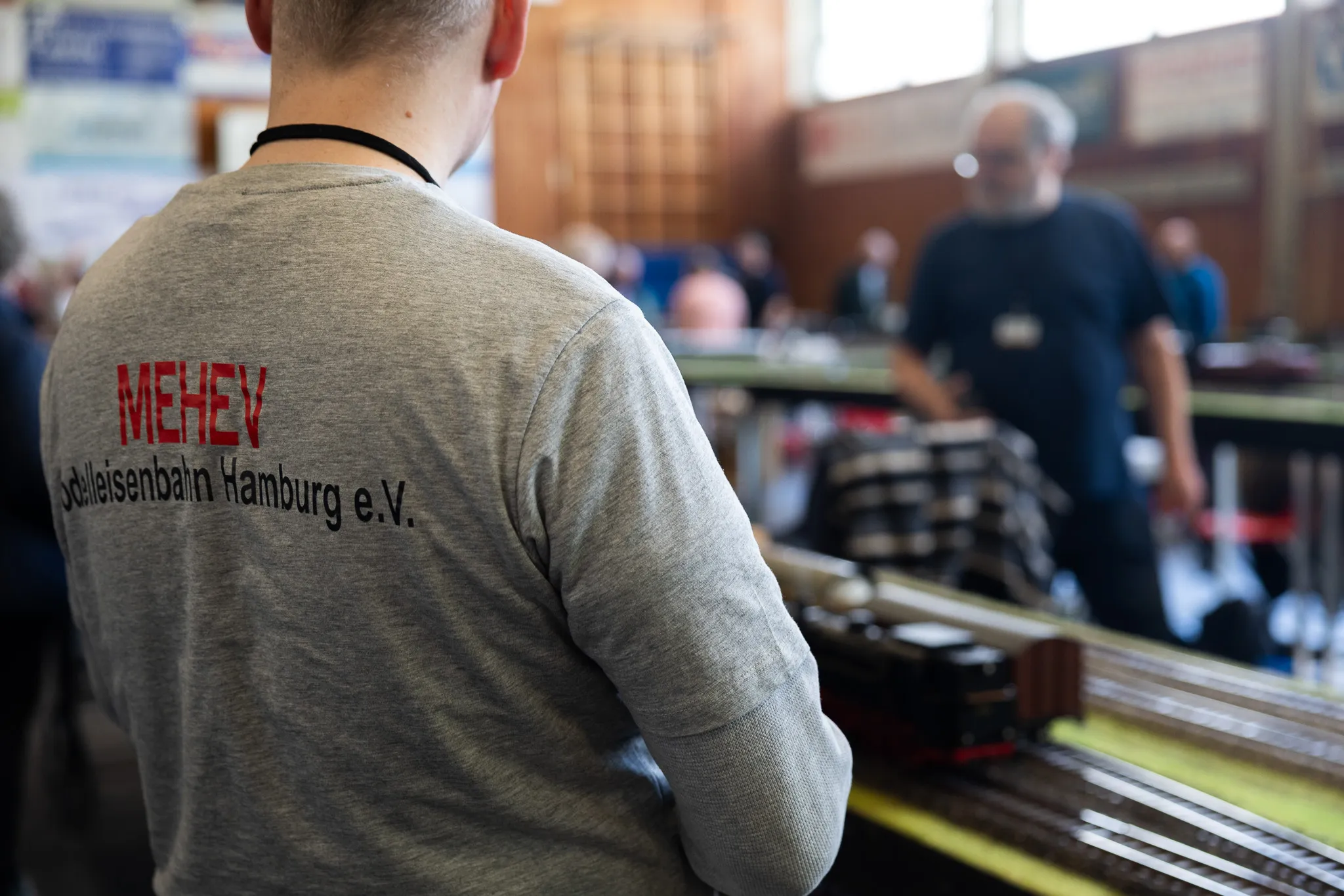 Rückenansicht einer Person mit dem Aufdruck „MEHEV Modelleisenbahn Hamburg e.V.“ auf dem grauen Pullover, im Hintergrund eine Modellbahnanlage und weitere Besucher.