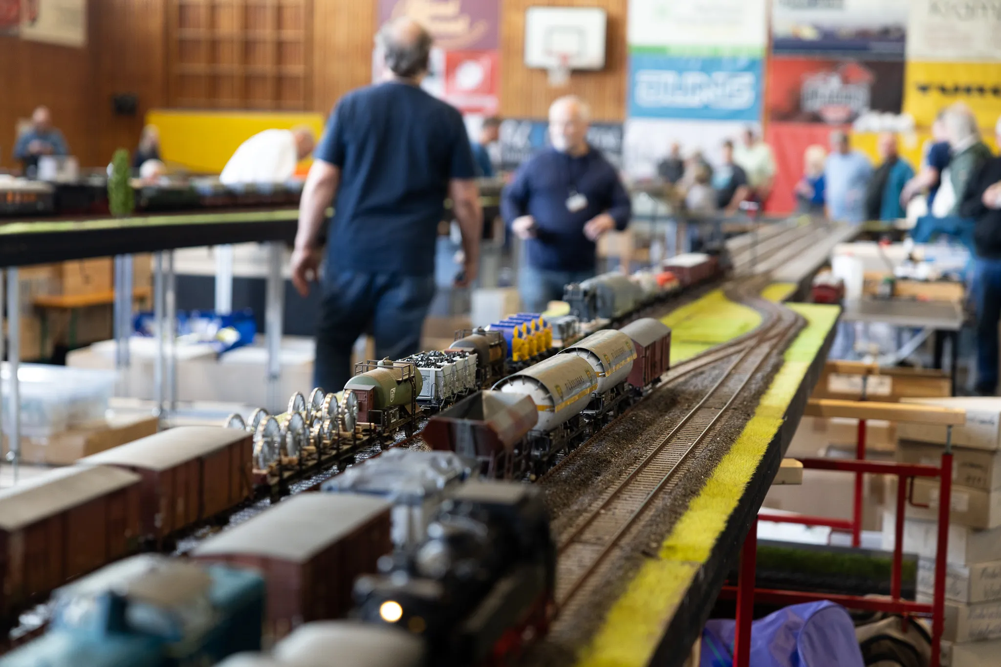 Eine detaillierte Modelleisenbahnanlage mit mehreren Waggons auf Gleisen steht im Fokus, während im Hintergrund Besucher eine Ausstellungshalle betreten.