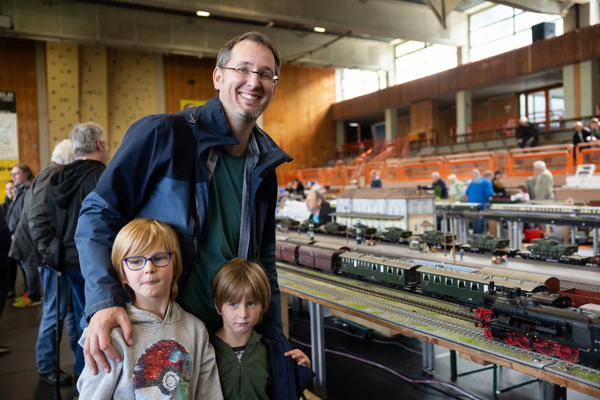 Ein Mann mit zwei Kindern steht vor einer detailreichen Modelleisenbahnanlage in einer Turnhalle. Im Hintergrund sind weitere Besucher und eine Zuschauertribüne zu sehen.