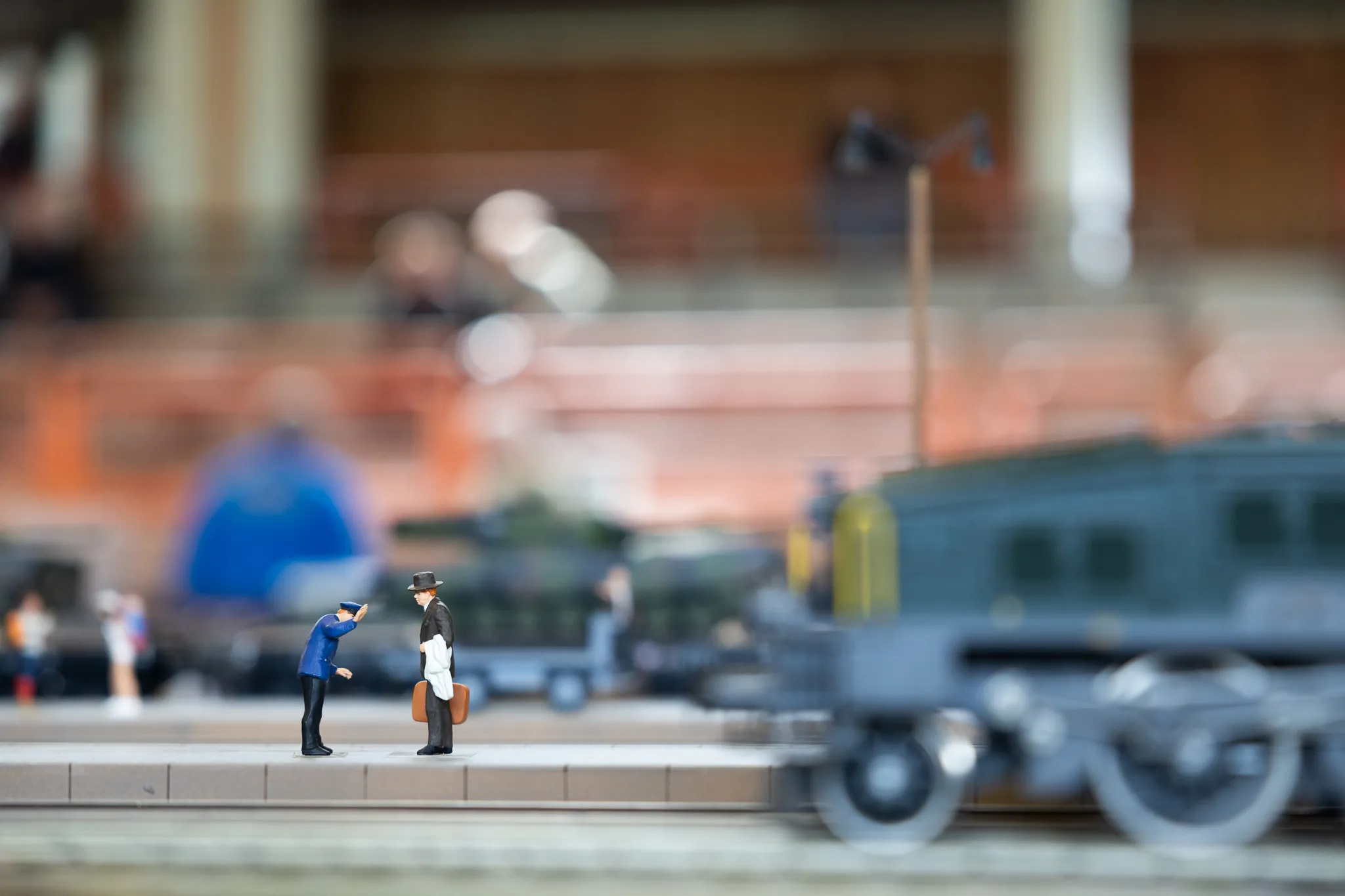 Zwei Miniaturfiguren stehen an einem Bahnsteig neben einer Dampflokomotive, im Hintergrund verschwommene weitere Figuren und eine Halle.