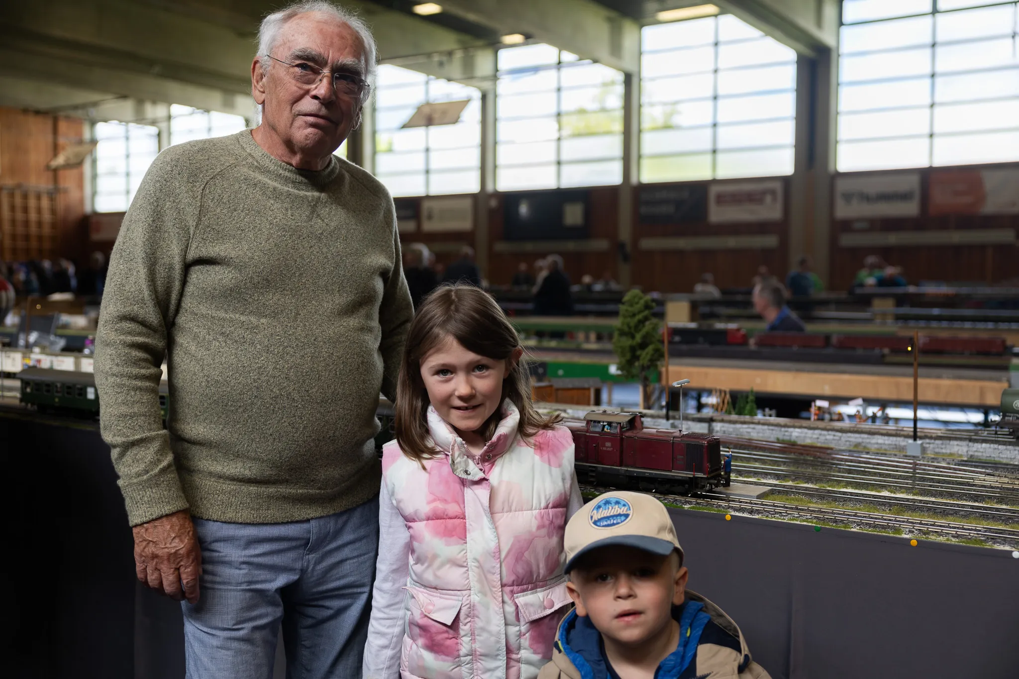 Ein älterer Mann mit zwei Kindern vor einer detaillierten Modelleisenbahnanlage in einer hellen Halle.