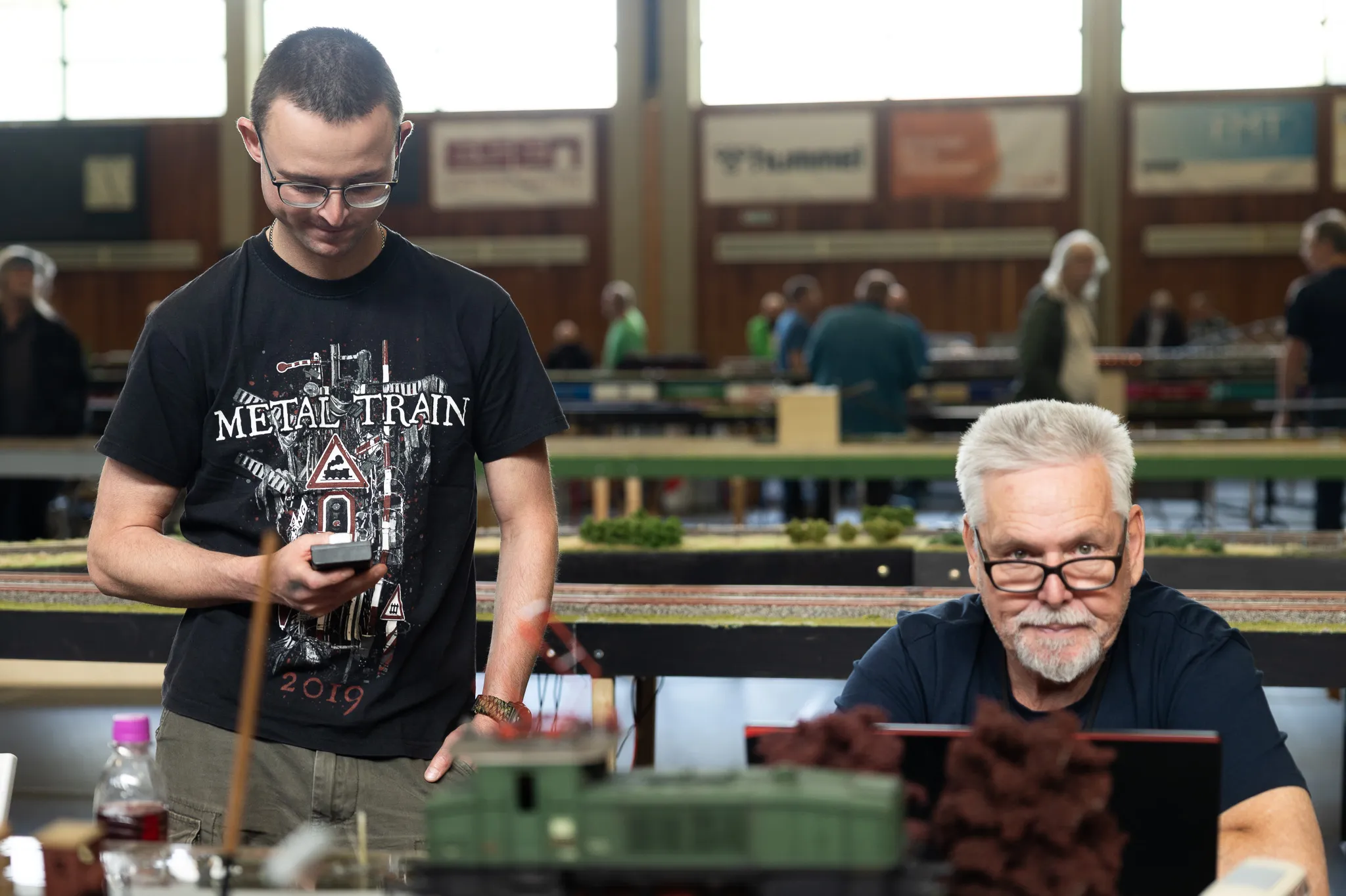 Zwei Männer betrachten eine detailreiche Modelleisenbahnanlage auf Tischen in einer hellen Halle. Der jüngere trägt ein T-Shirt mit dem Aufdruck „Metal Train“.