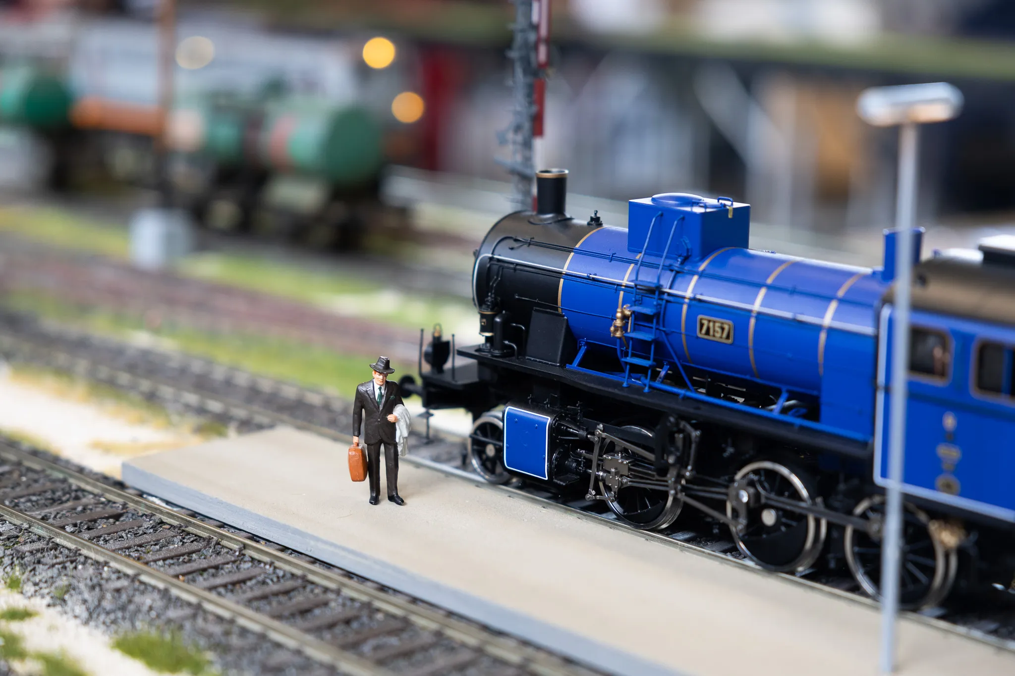 Blaue Dampflok steht auf Gleisen einer Modellbahn neben einem Mann mit Koffer.