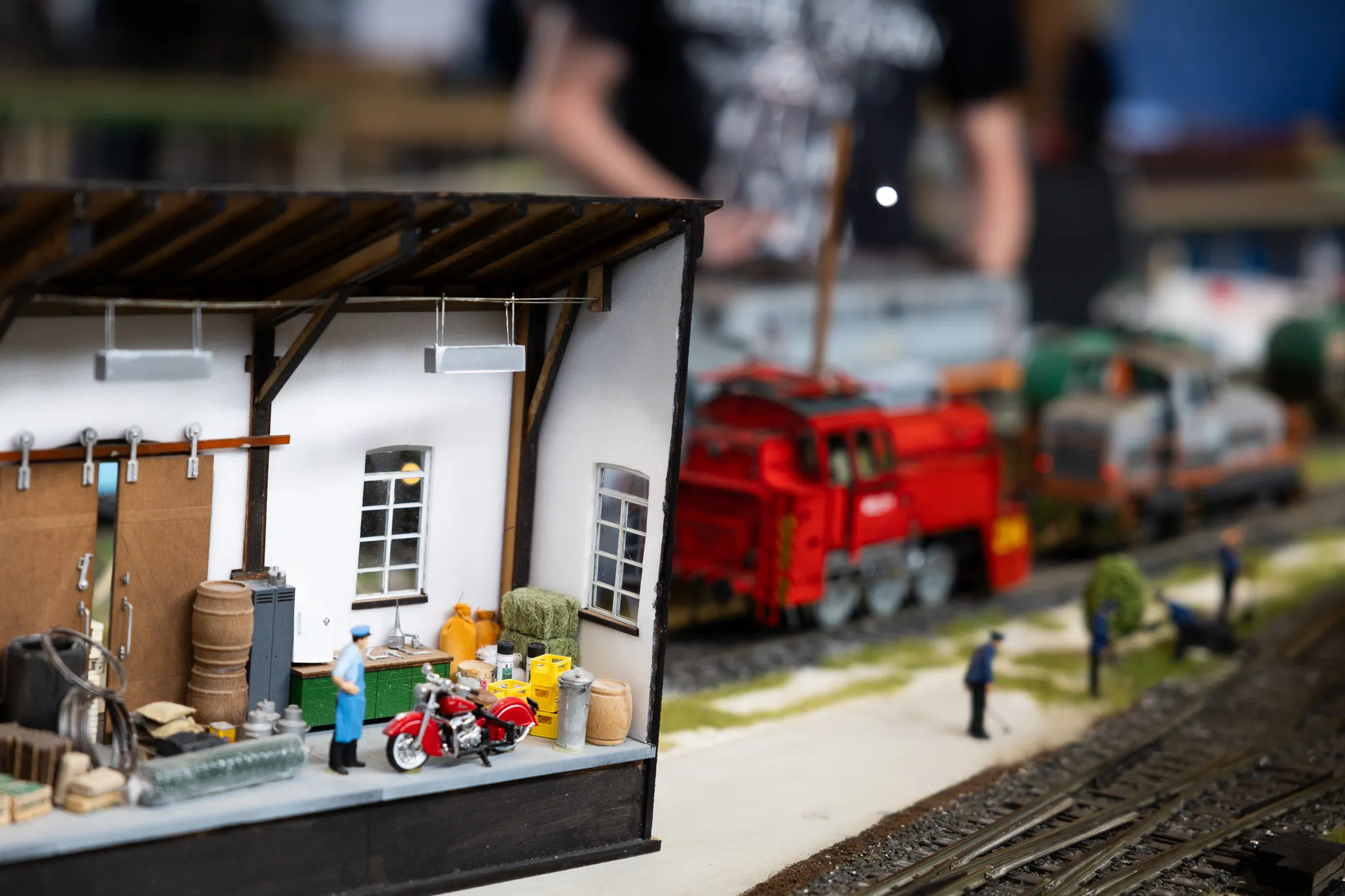 Detailansicht einer Modellbahnanlage mit einem Gebäude, einer roten Lokomotive und kleinen Figuren, die eine Szene darstellen.