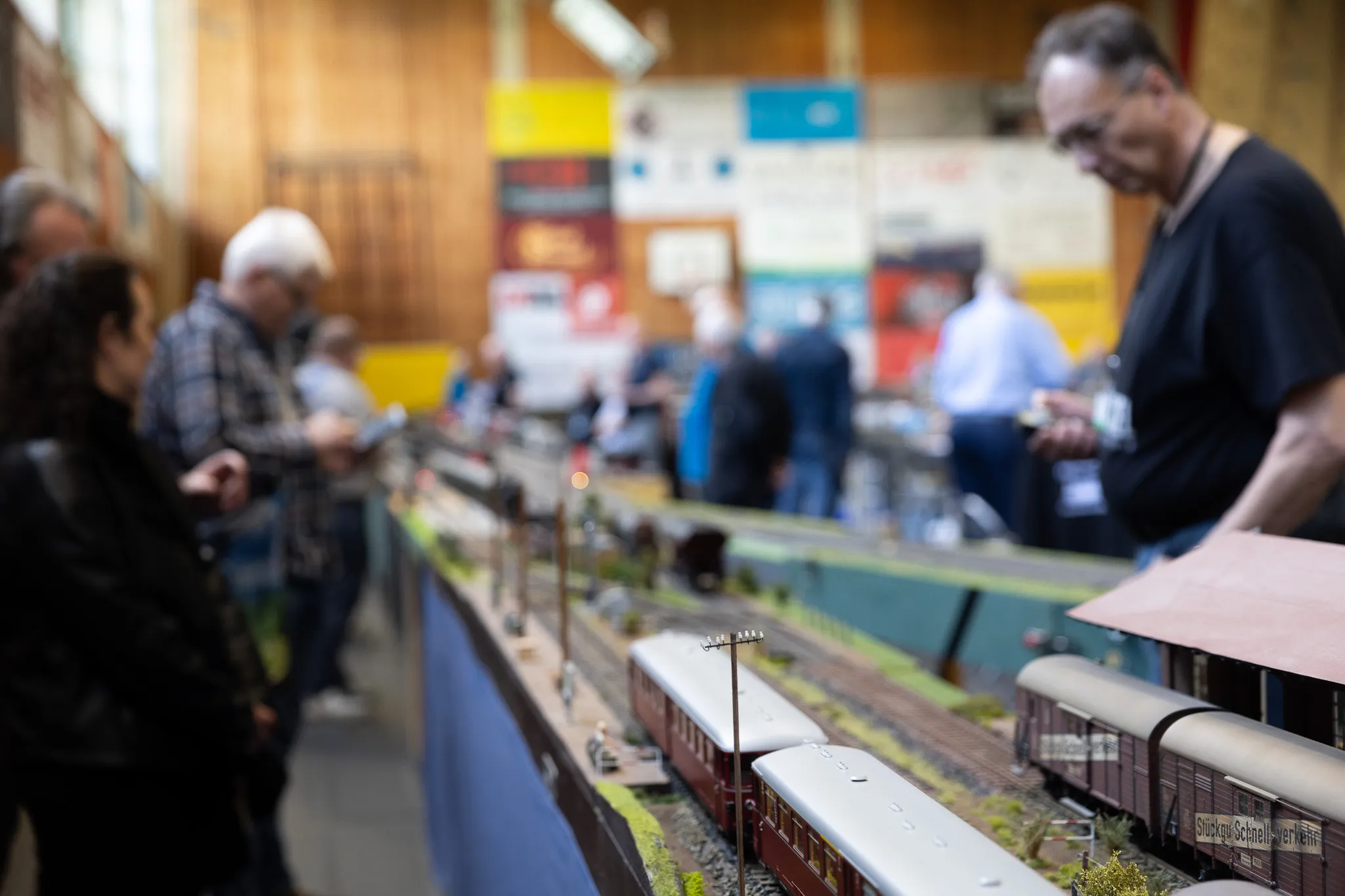 Modellbahn-Anlage mit mehreren Zugwaggons im Vordergrund, im Hintergrund Besucher einer Ausstellung in einer Turnhalle.
