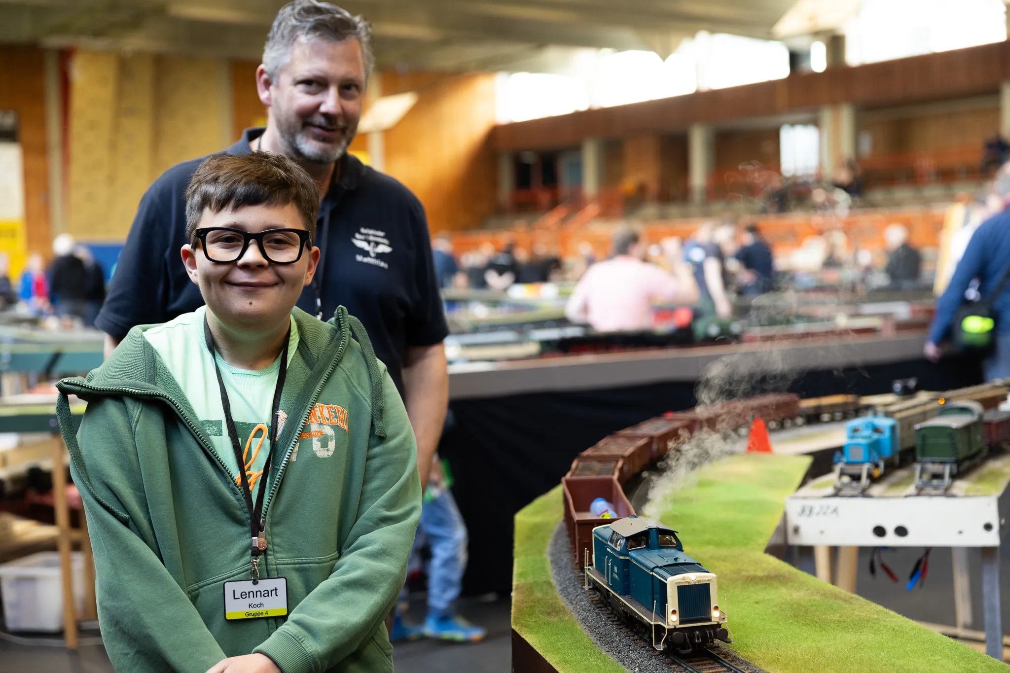 Ein Junge mit Brille und ein Mann stehen vor einer Miniatur-Eisenbahnanlage mit fahrenden Zügen und Landschaftselementen.