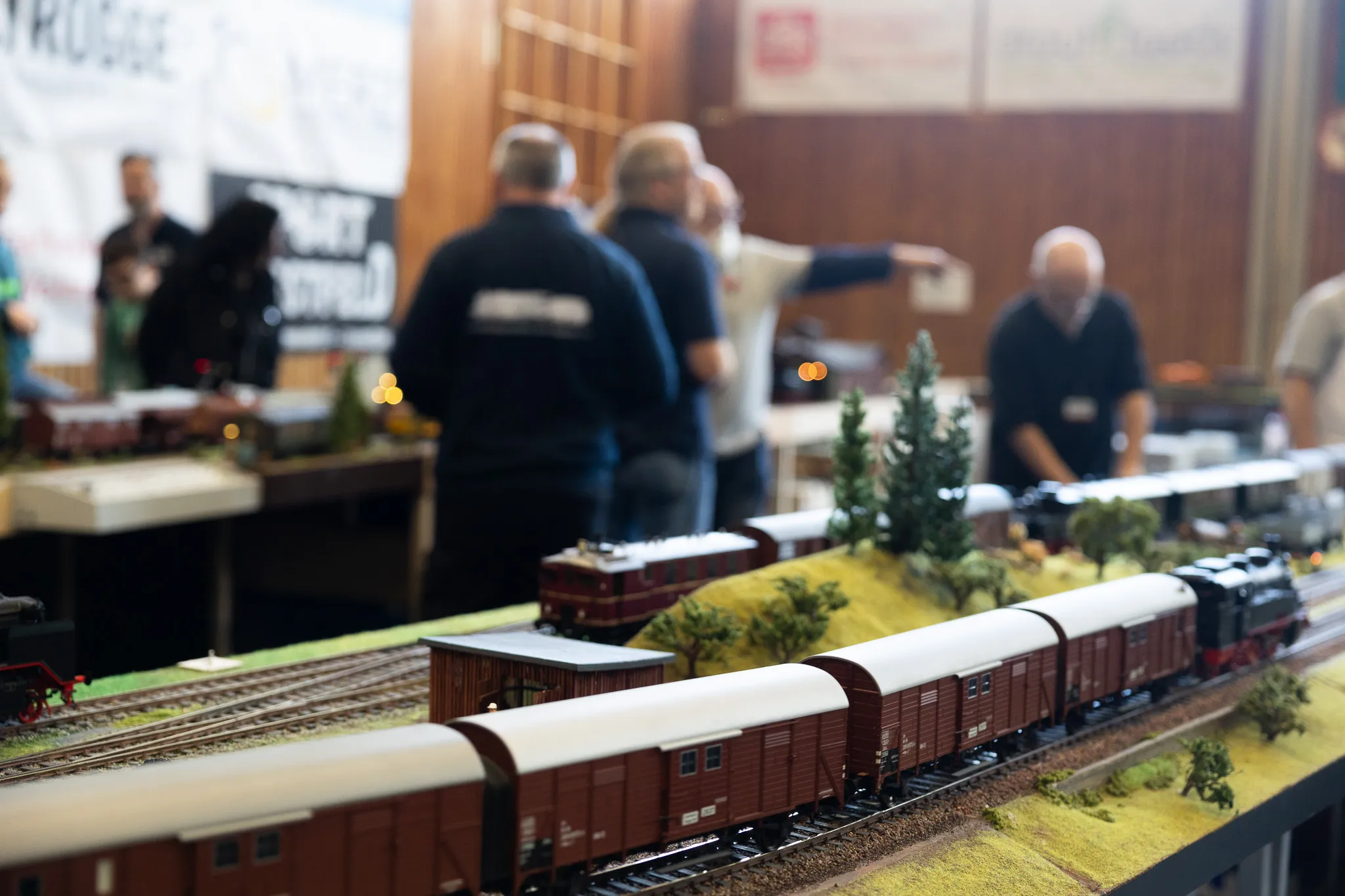 Nahaufnahme einer detaillierten Modelleisenbahnanlage mit mehreren Waggons auf Gleisen, im Hintergrund verschwommene Besucher einer Ausstellung.