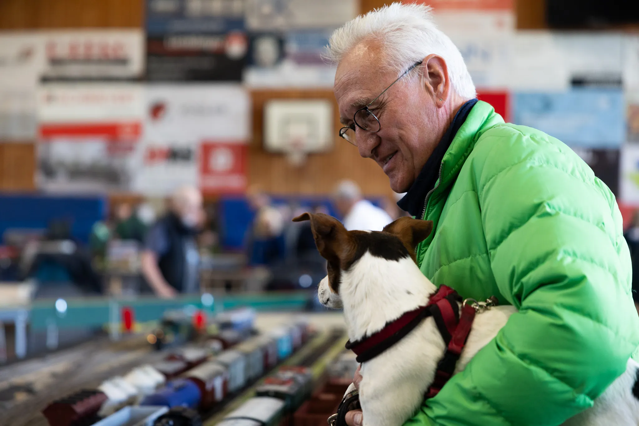 Ein älterer Mann mit weißem Haar und grüner Jacke blickt auf einen kleinen, weiß-schwarzen Hund, der auf einem Tisch vor ihm steht. Im Hintergrund sind weitere Besucher und eine Modelleisenbahnanlage zu sehen.