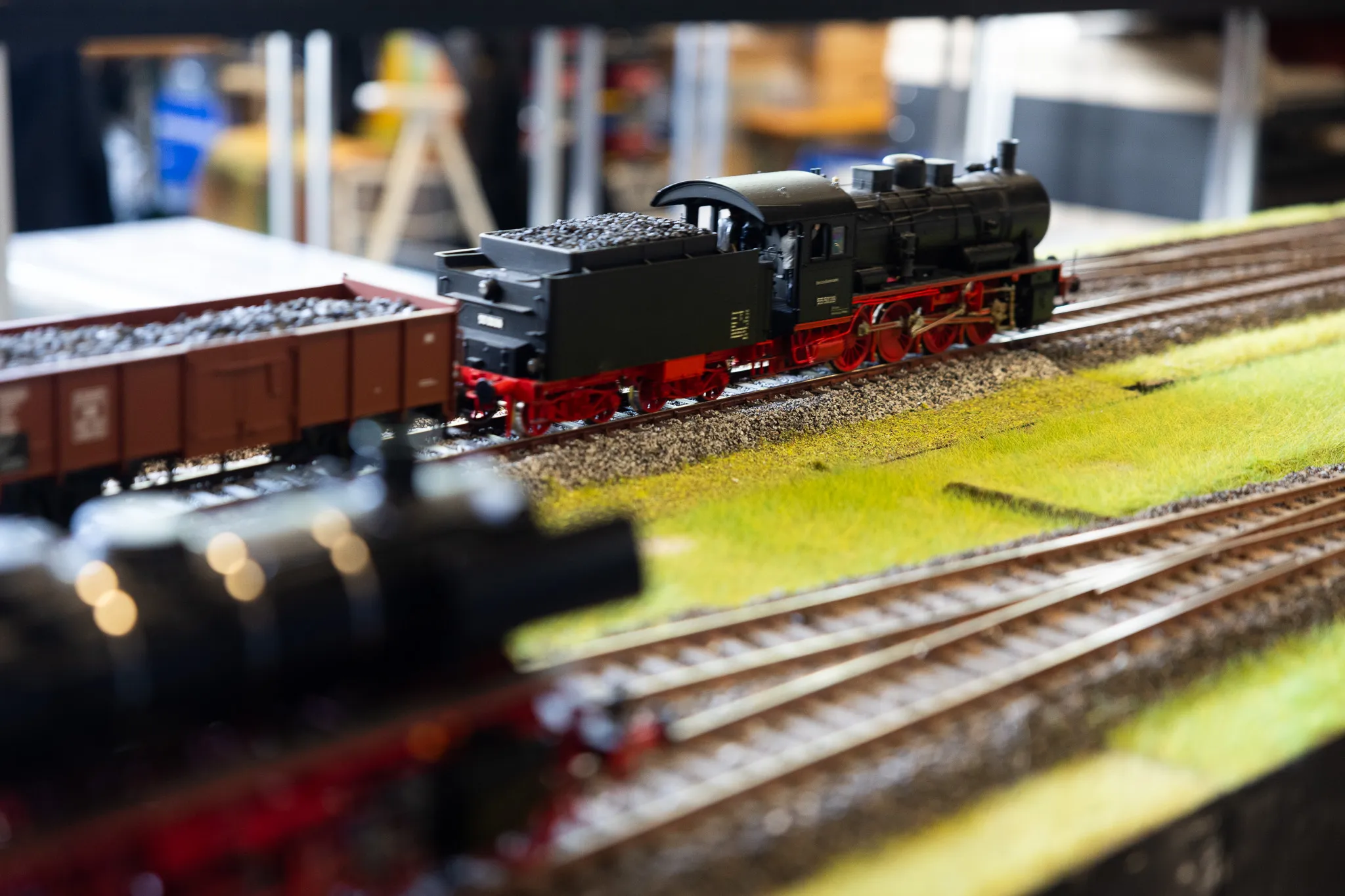 Schwarze Dampflokomotive mit roten Rädern und Waggons auf einer Modellbahn-Landschaft mit grünem Rasen.