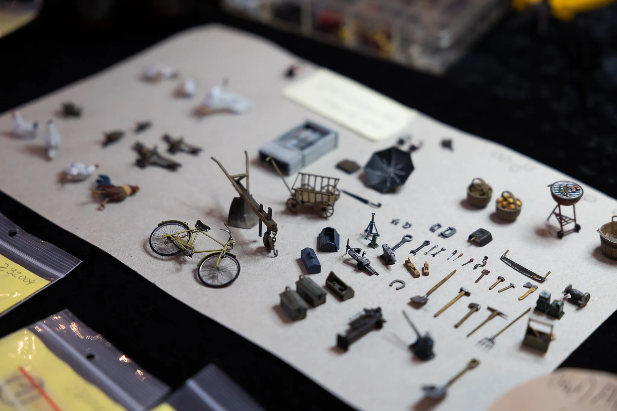 Detailaufnahme einer Anordnung von Miniaturfiguren und Objekten auf einer grauen Oberfläche, darunter Fahrräder, Möbel und Werkzeuge, vermutlich Teil einer Modellbahnanlage.