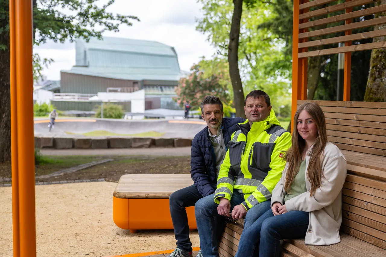 Drei Personen sitzen auf einer Holzbank vor einem Skatepark und einem modernen Gebäude. Von links sind Marcus Müller, Stefan Tackmann und Nora Nieswand zu sehen.