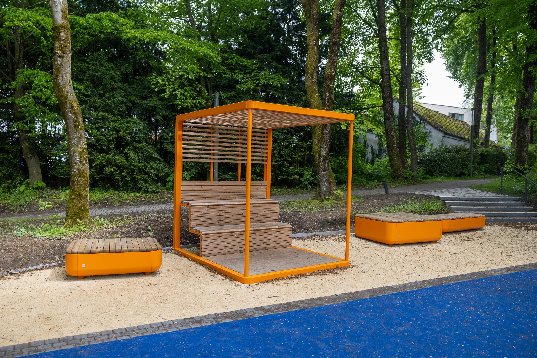 Orangefarbener Pavillon mit Holzbänken auf einem Kiesplatz, im Hintergrund Bäume und ein Wohnhaus.