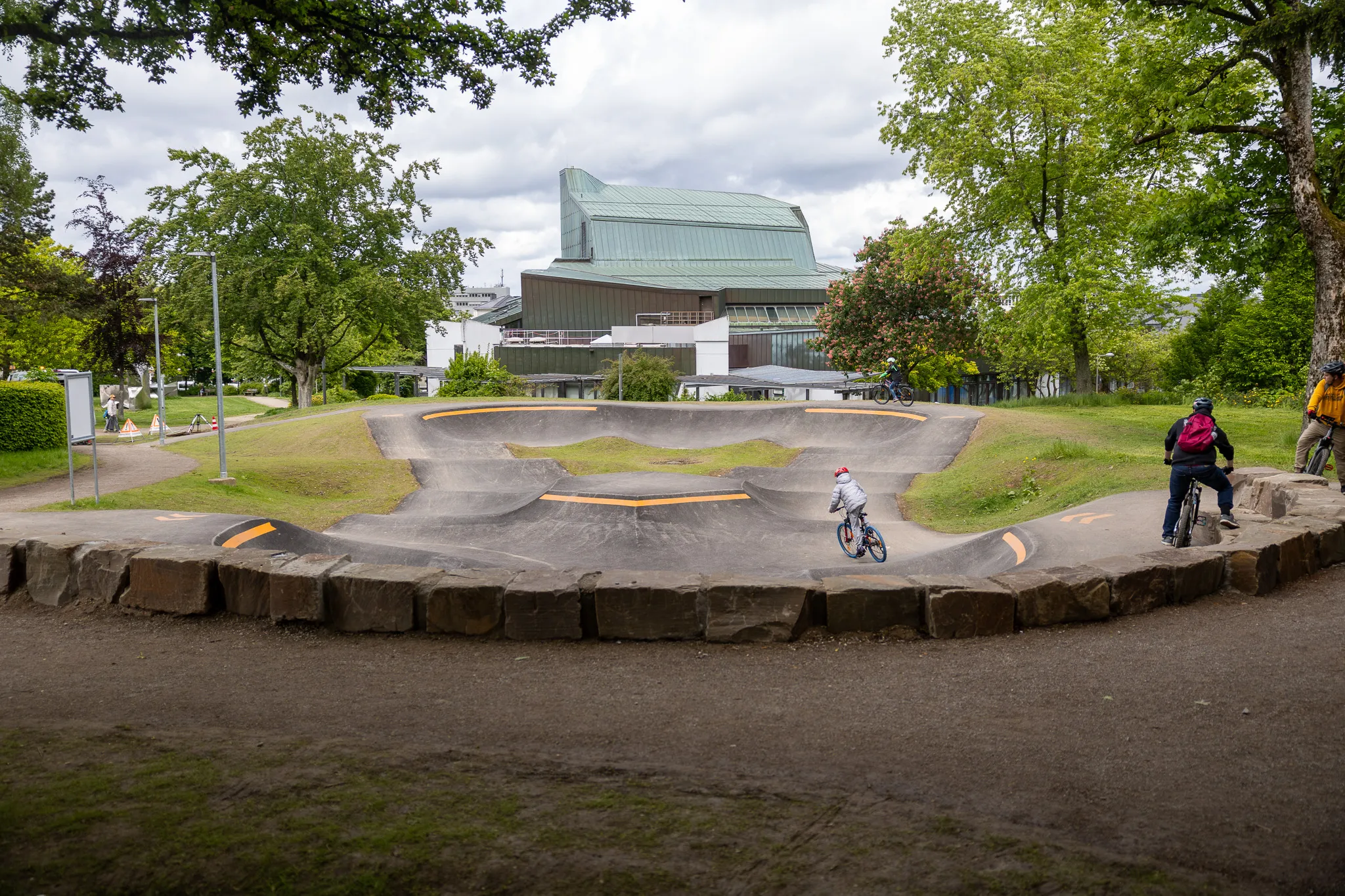 Zwei Kinder fahren auf einem Pumptrack im Lüdenscheider Stadtgarten, im Hintergrund ein modernes Gebäude.