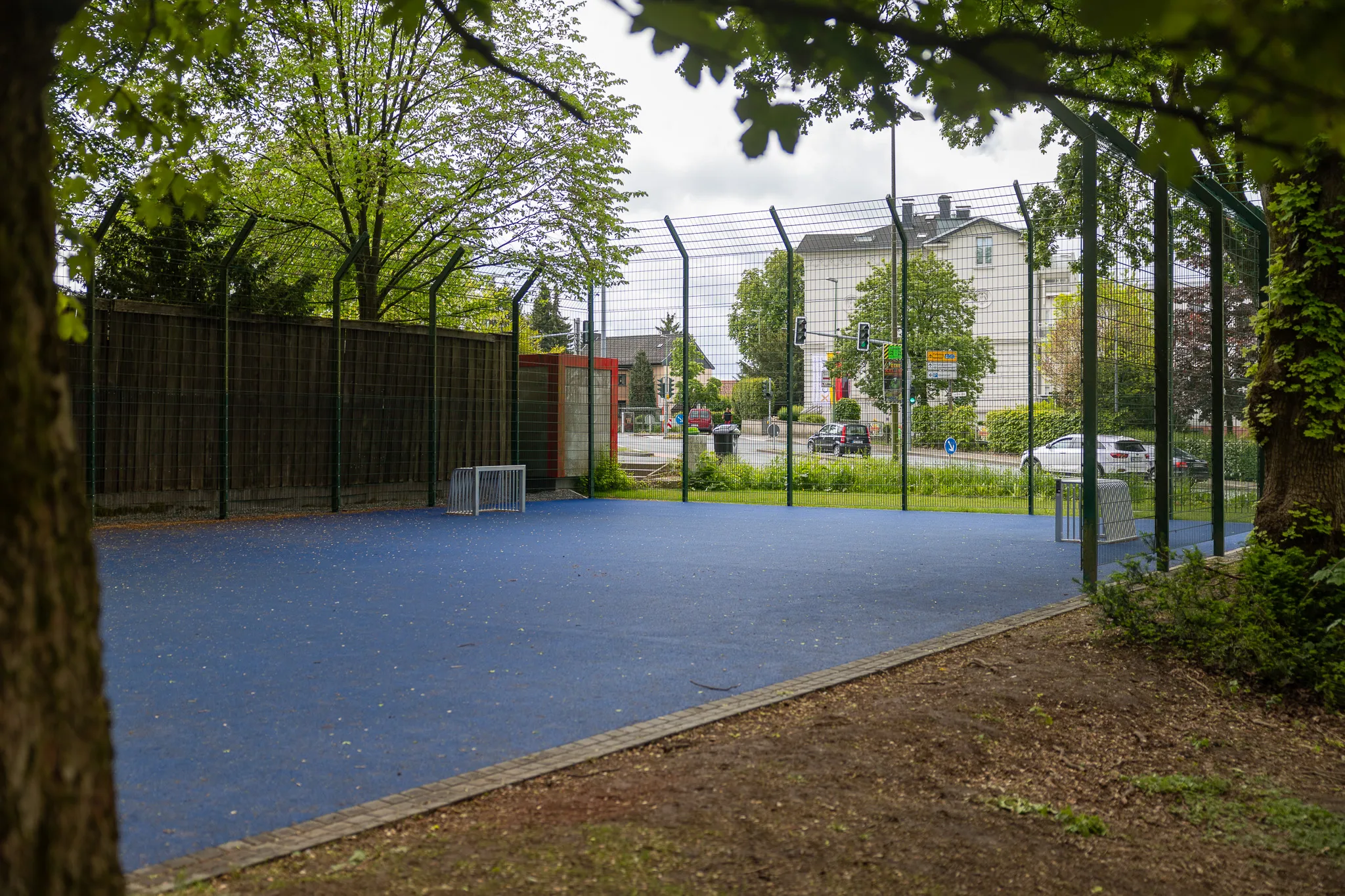 Blauer Sportplatz mit Maschendrahtzaun und Bäumen im Hintergrund, daneben ein roter Schrank und im Hintergrund Gebäude und Straßenverkehr.