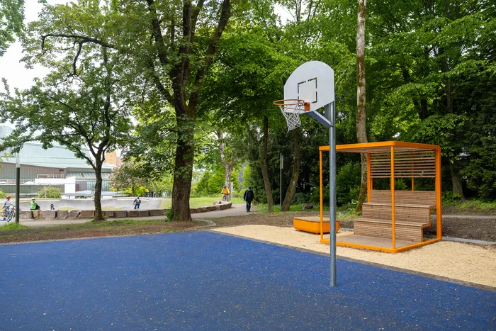 Basketballkorb und orangefarbene Sitzgelegenheit neben einem blauen Basketballfeld im Lüdenscheider Stadtgarten. Im Hintergrund ein Pumptrack und ein Gebäude.