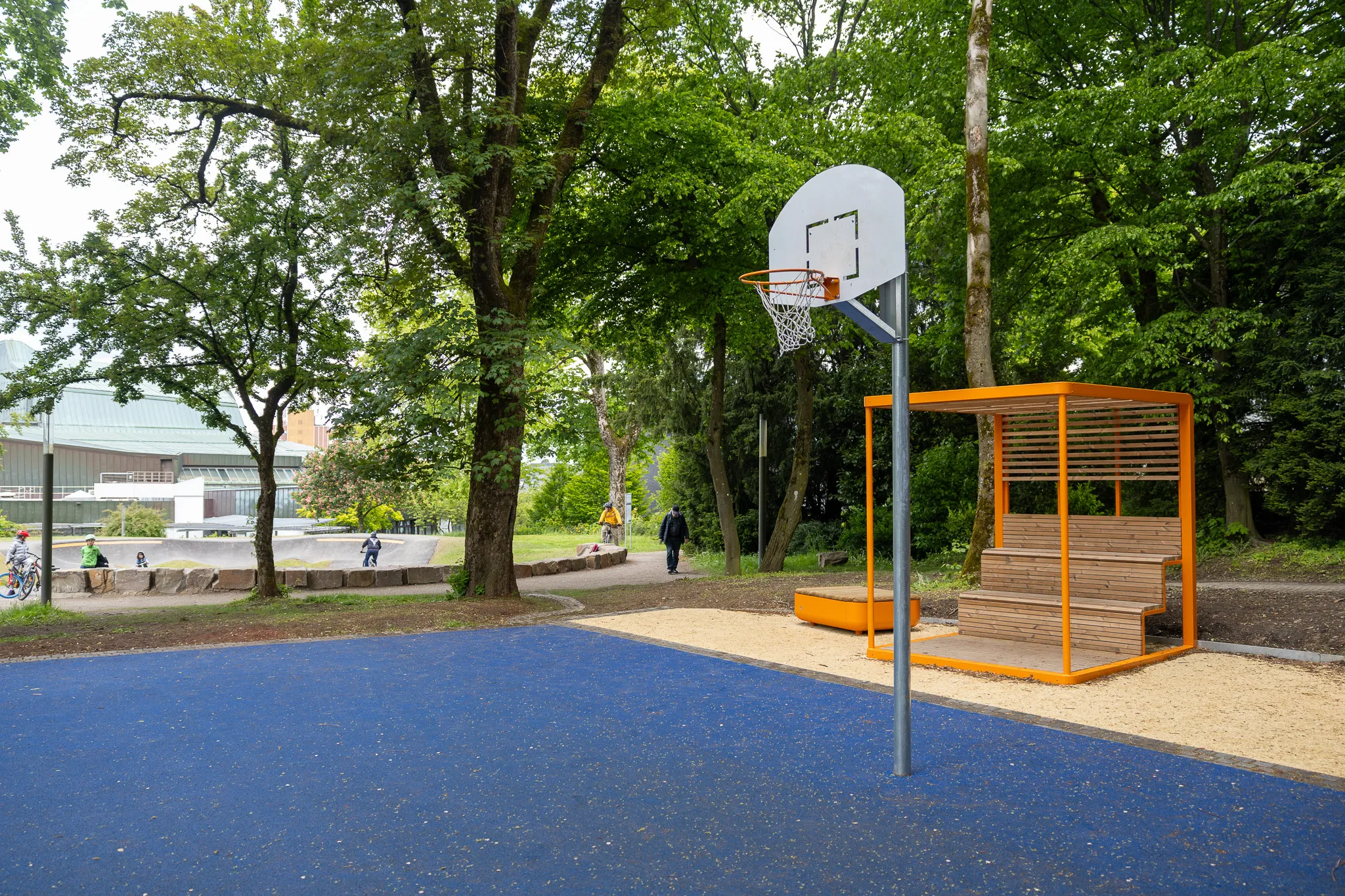 Basketballkorb und orangefarbene Sitzgelegenheit neben einem blauen Basketballfeld im Lüdenscheider Stadtgarten. Im Hintergrund ein Pumptrack und ein Gebäude.