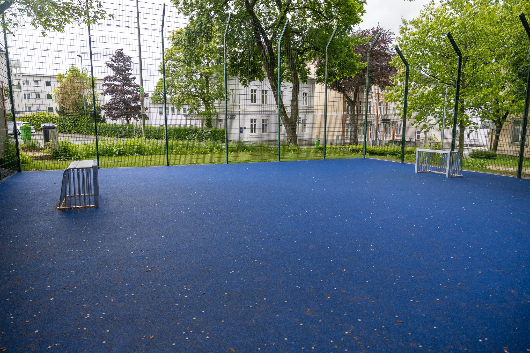 Blauer Sportplatz mit Banden und zwei kleinen Hockeytoren, umgeben von Bäumen und Büschen im Lüdenscheider Stadtgarten.