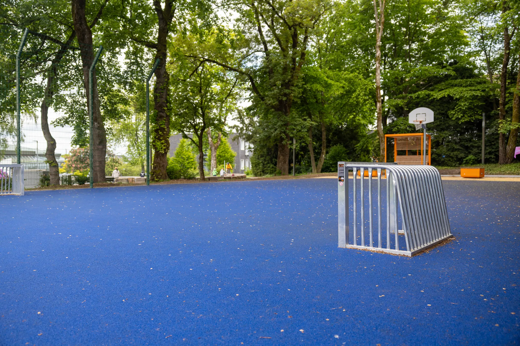 Blauer Sportplatz mit Metallgeländer im Vordergrund und Basketballkorb sowie Bäumen im Hintergrund.