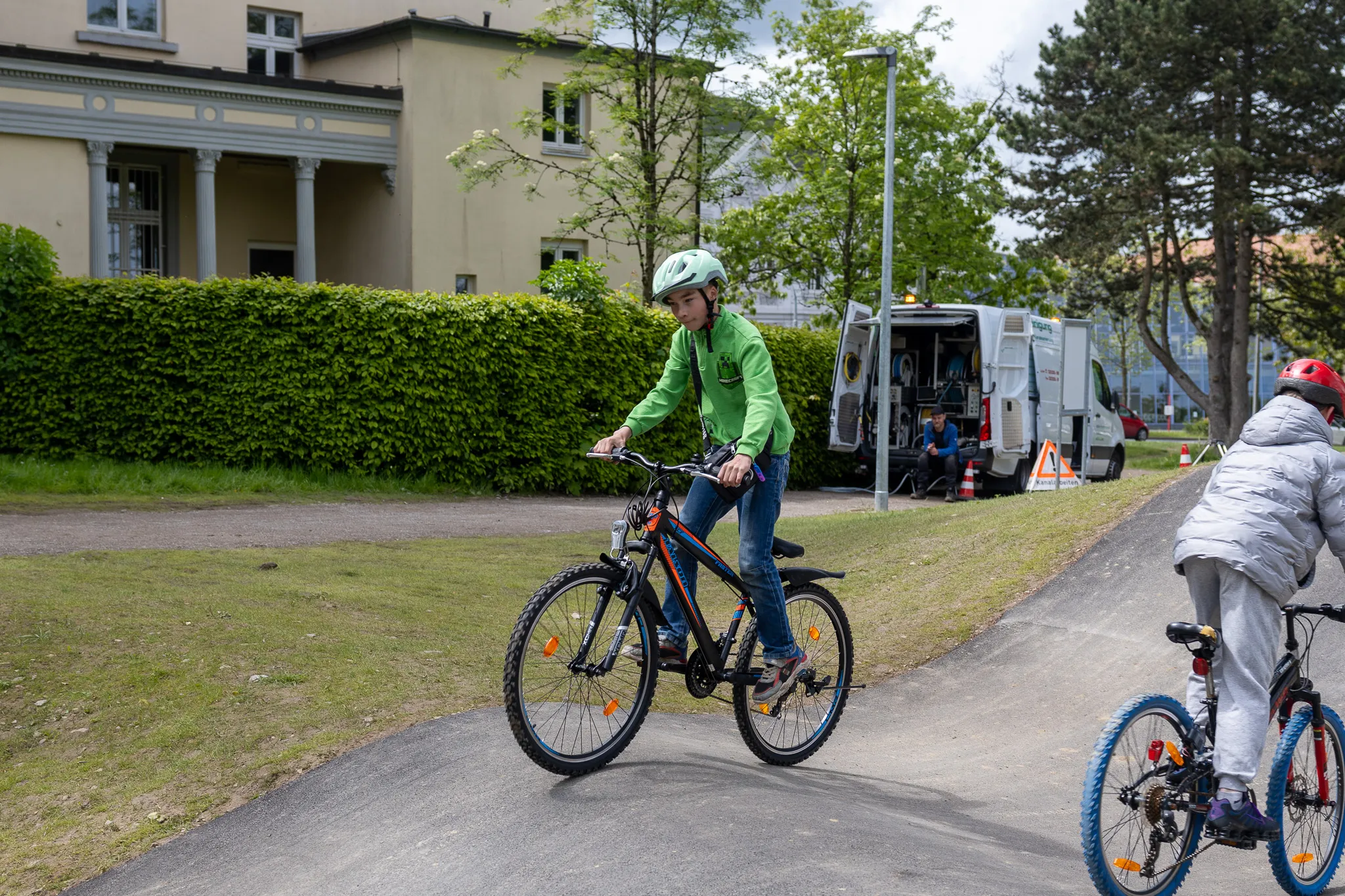 Zwei Kinder fahren mit Fahrrädern auf einem asphaltierten Weg im Lüdenscheider Stadtgarten, im Hintergrund ein Gebäude und ein Transporter.
