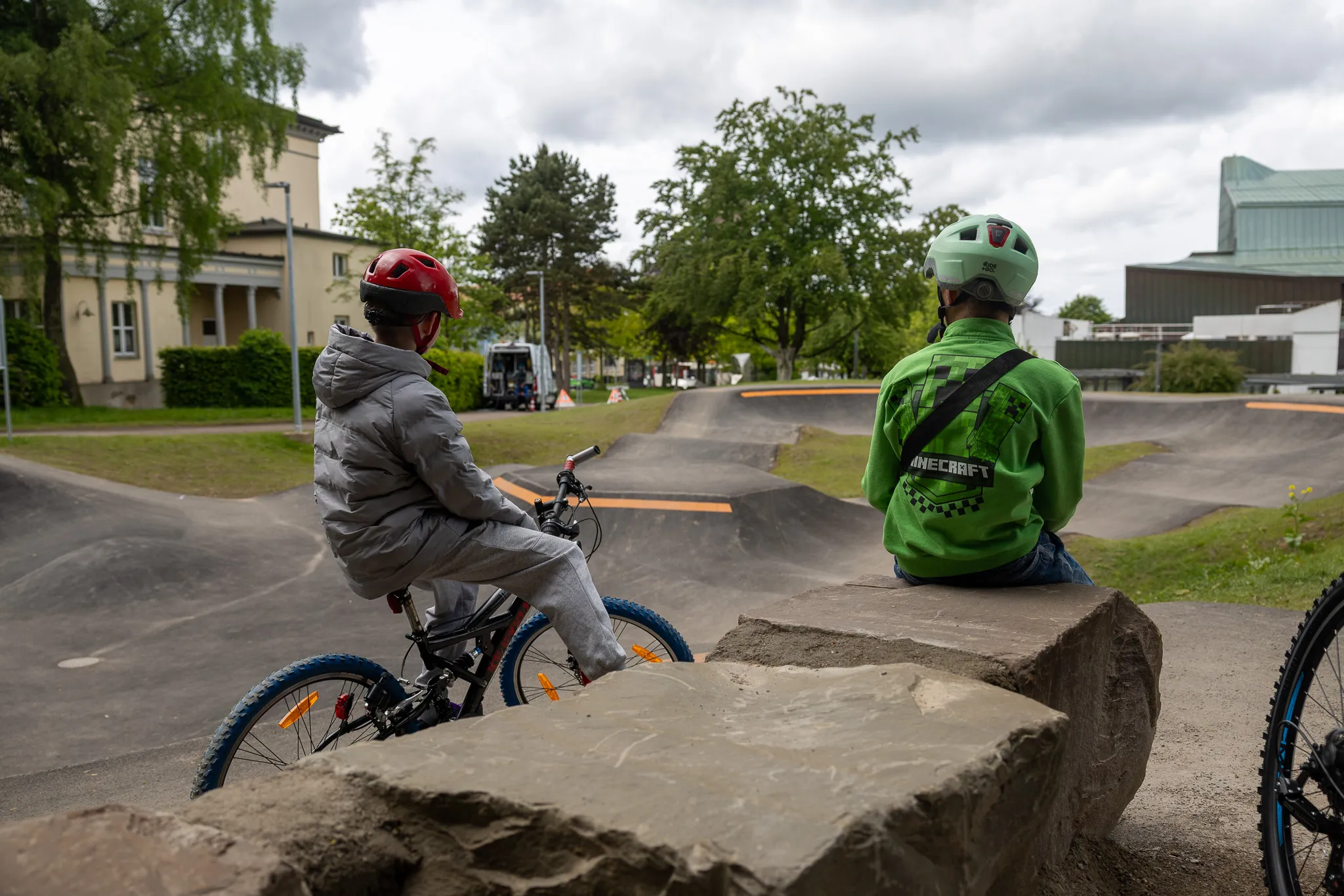 Zwei Kinder mit Fahrrädern und Helmen sitzen auf einer erhöhten Plattform eines Pumptracks im Stadtgarten, während ein dritter Radfahrer im Hintergrund fährt.
