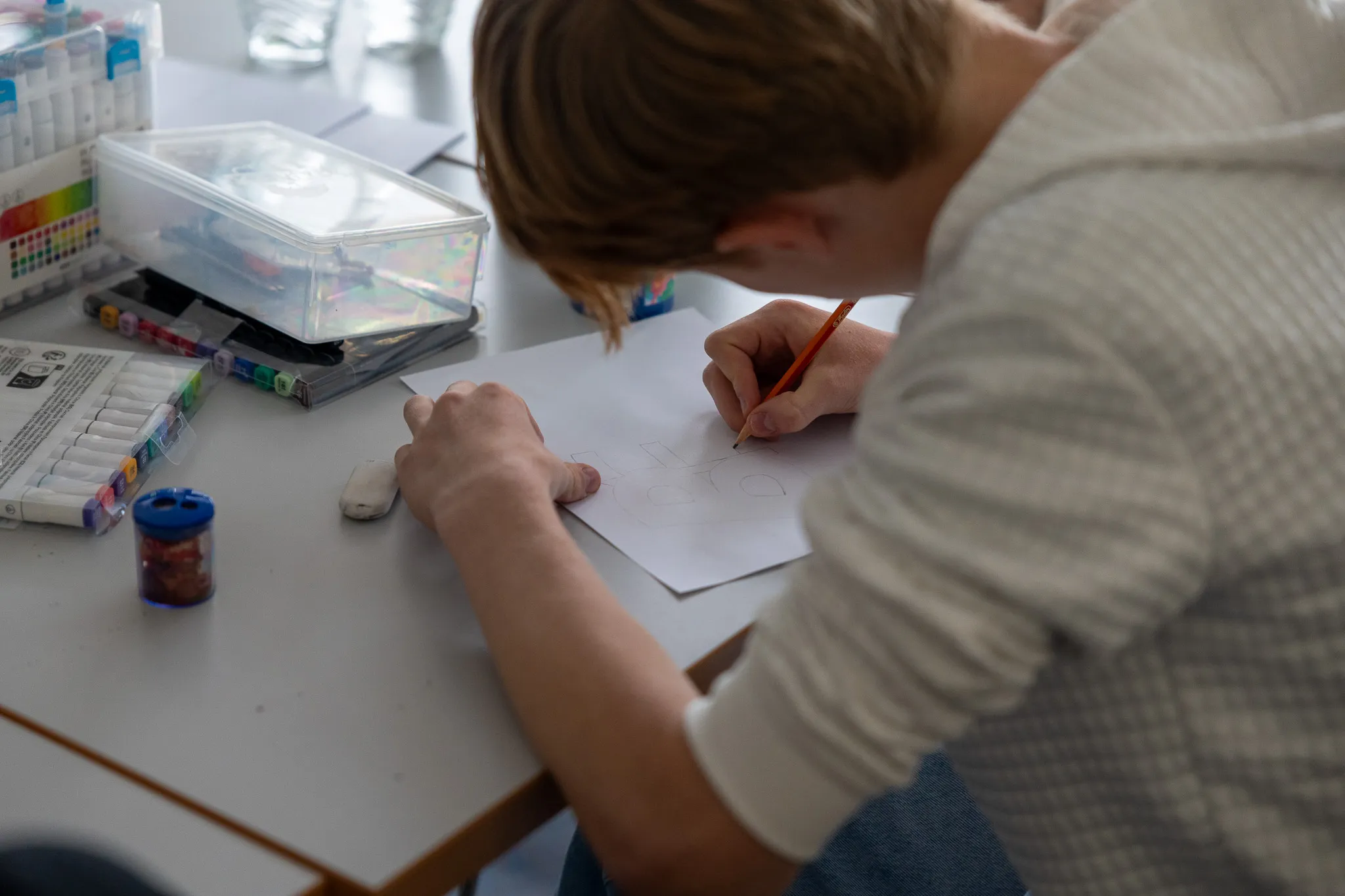 Eine Person mit kurz geschnittenem braunem Haar zeichnet mit einem orangefarbenen Stift auf ein weißes Blatt Papier. Im Hintergrund sind Farbstifte und andere Zeichenmaterialien auf einem Tisch zu sehen.