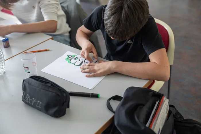 Ein Jugendlicher mit dunklem T-Shirt und kurzen Haaren konzentriert sich auf das Ausmalen einer Graffiti-Vorlage mit einem Filzstift an einem Tisch in einem Raum.