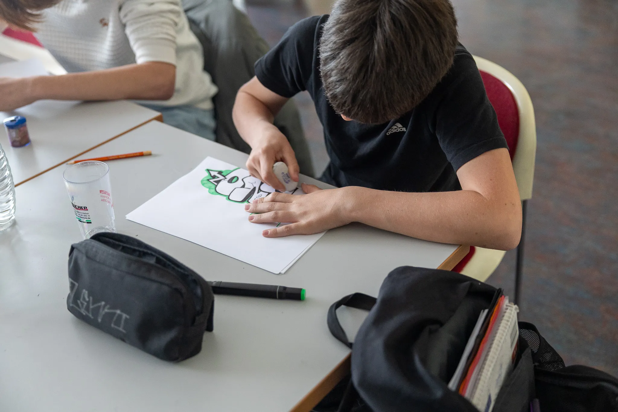 Ein Jugendlicher mit dunklem T-Shirt und kurzen Haaren konzentriert sich auf das Ausmalen einer Graffiti-Vorlage mit einem Filzstift an einem Tisch in einem Raum.