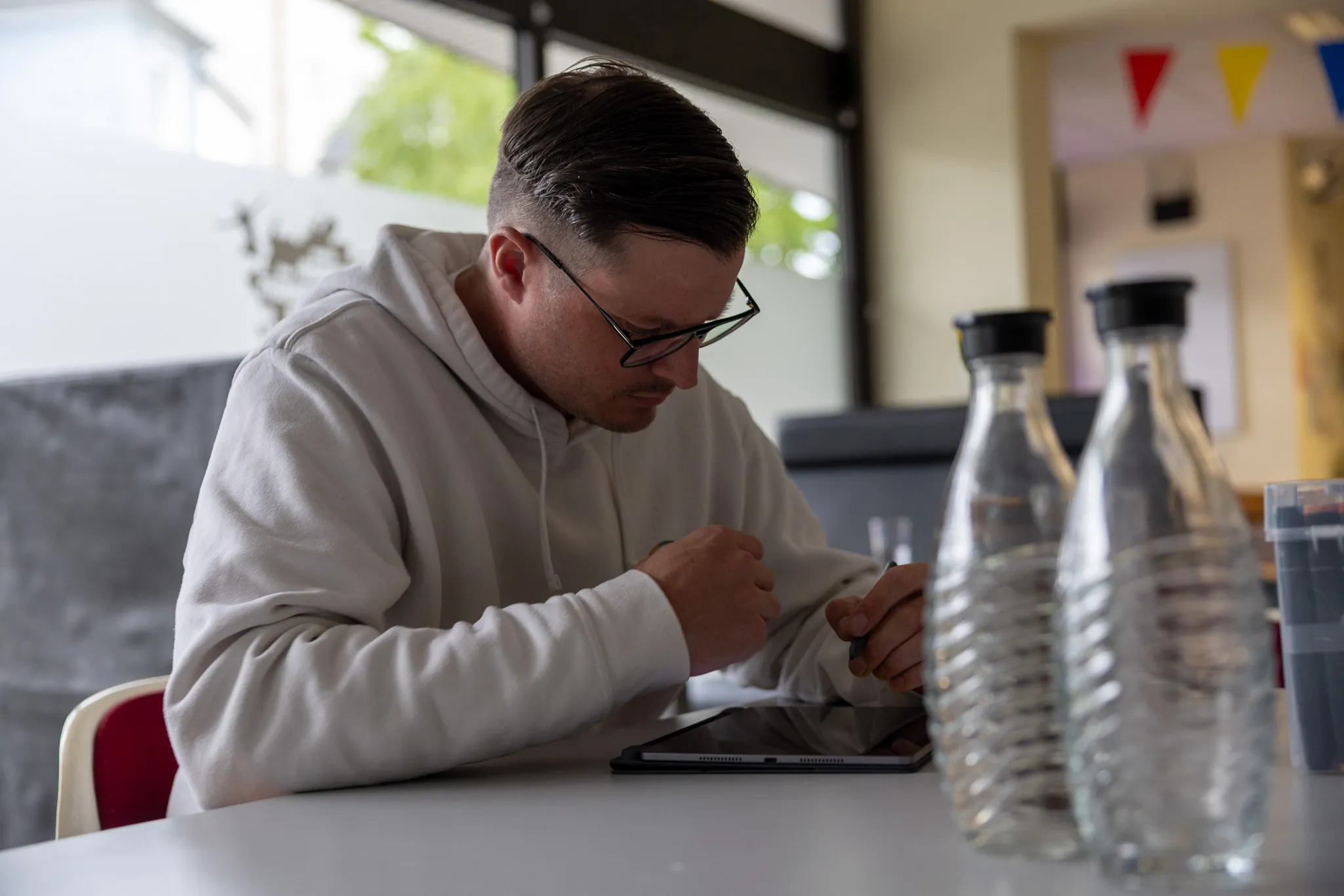 Ein Mann mit Brille und Kapuzenpulli zeichnet konzentriert mit einem Stift auf ein Tablet. Im Hintergrund sind bunte Wimpel und Wasserflaschen zu sehen.