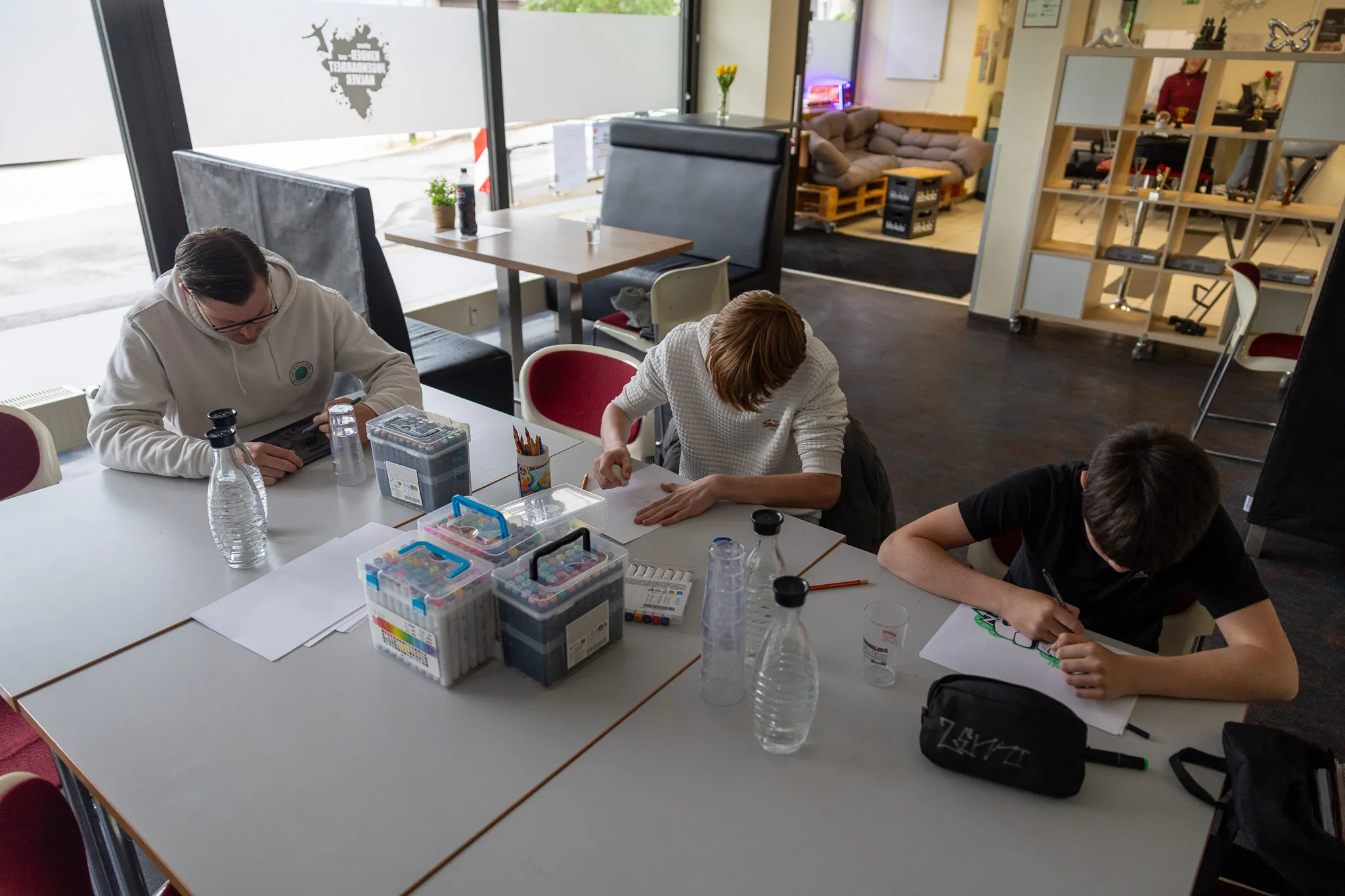 Drei Personen sitzen an einem Tisch und zeichnen mit Stiften und Papier. Im Hintergrund sind weitere Möbel und Personen in einem hellen Raum zu sehen.